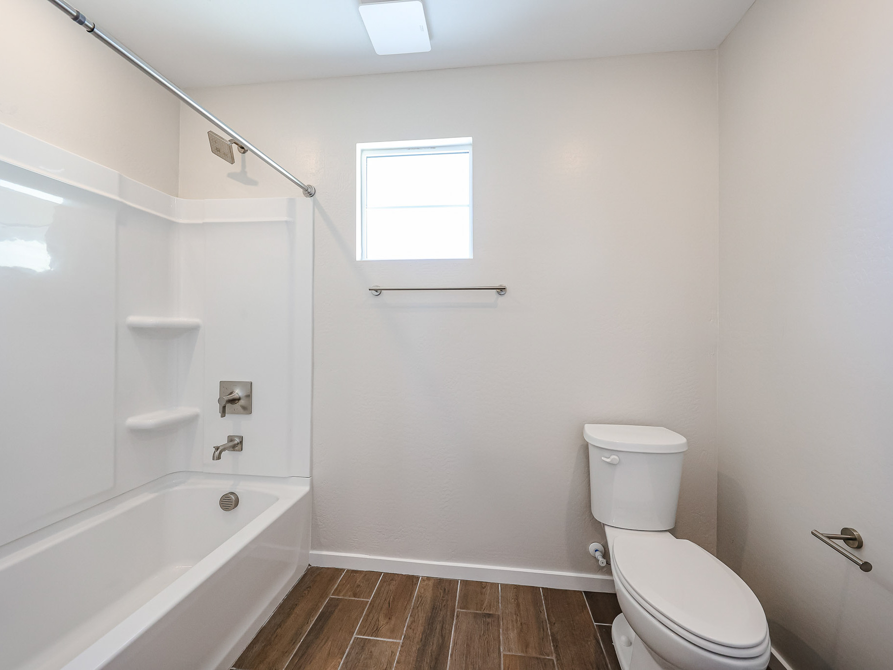 A bathroom with a white bathtub and shower, a white toilet, a towel bar, and wood-look tile flooring under a small window.