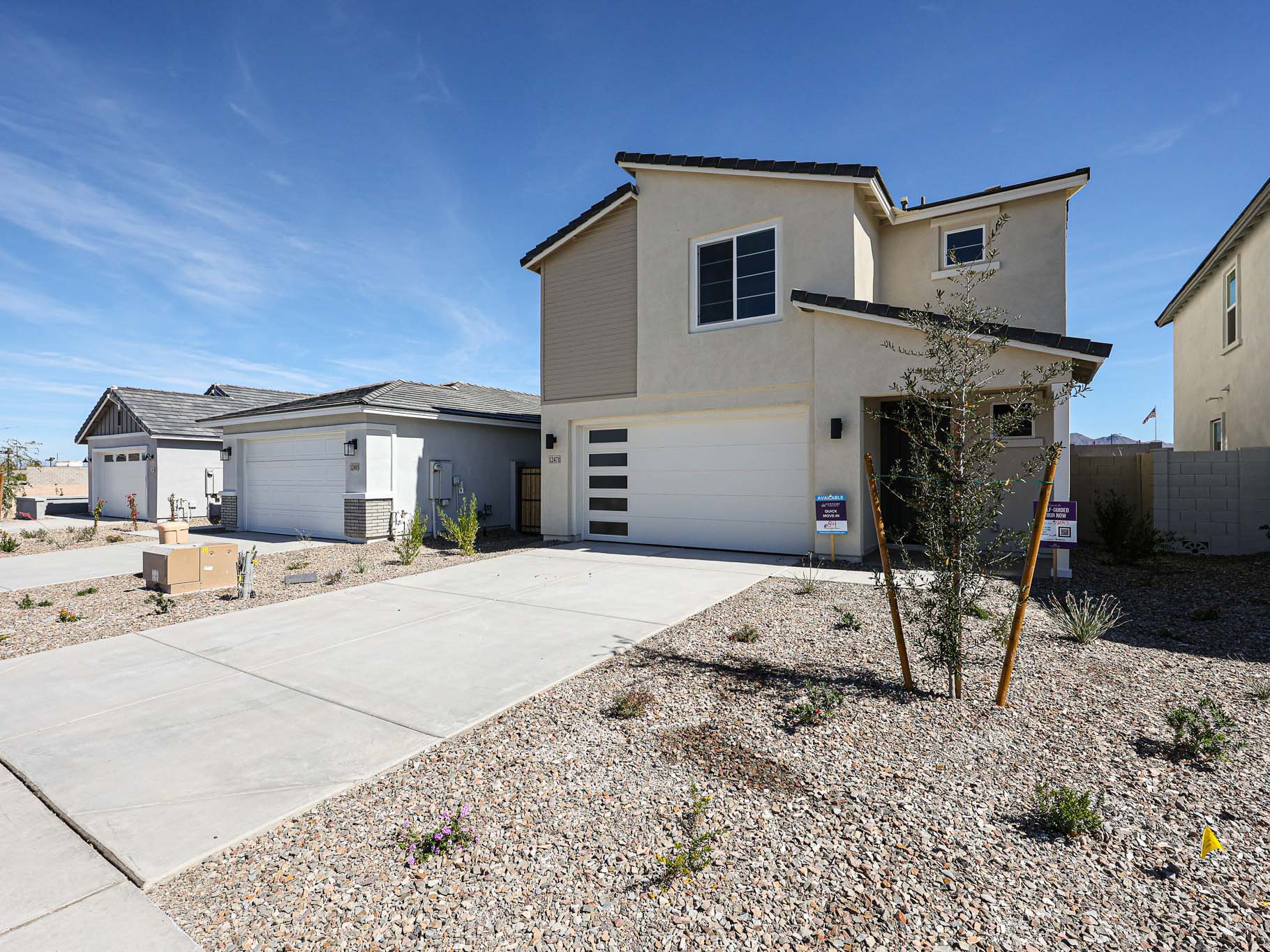 Two-story modern house with a white garage door, minimal landscaping, and a "For Sale" sign in the gravel front yard under a clear blue sky.