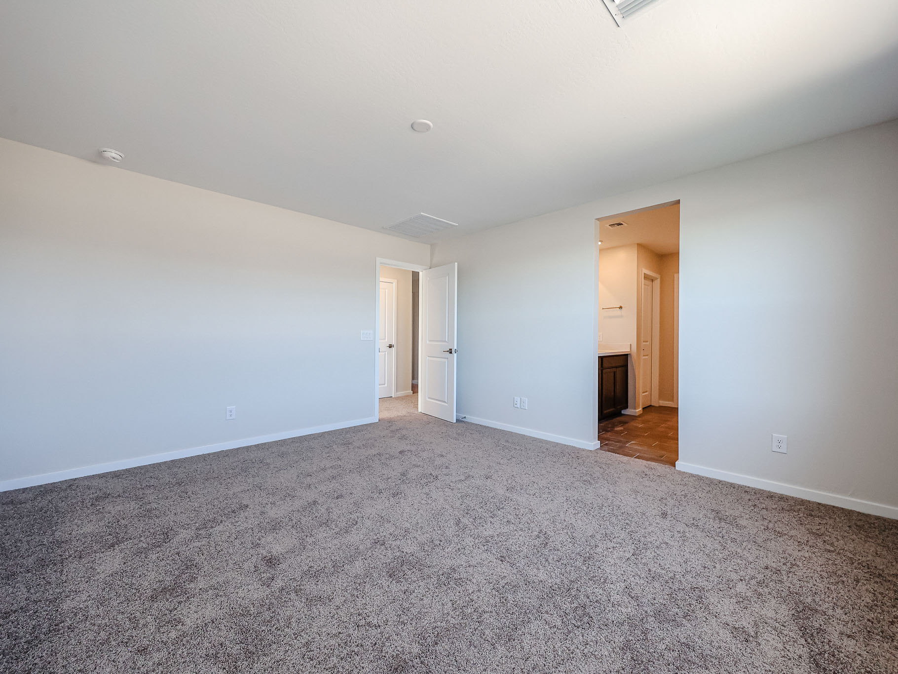 Empty room with beige carpet, white walls, an open door leading to a hallway, and an open doorway leading to a bathroom with tile flooring.