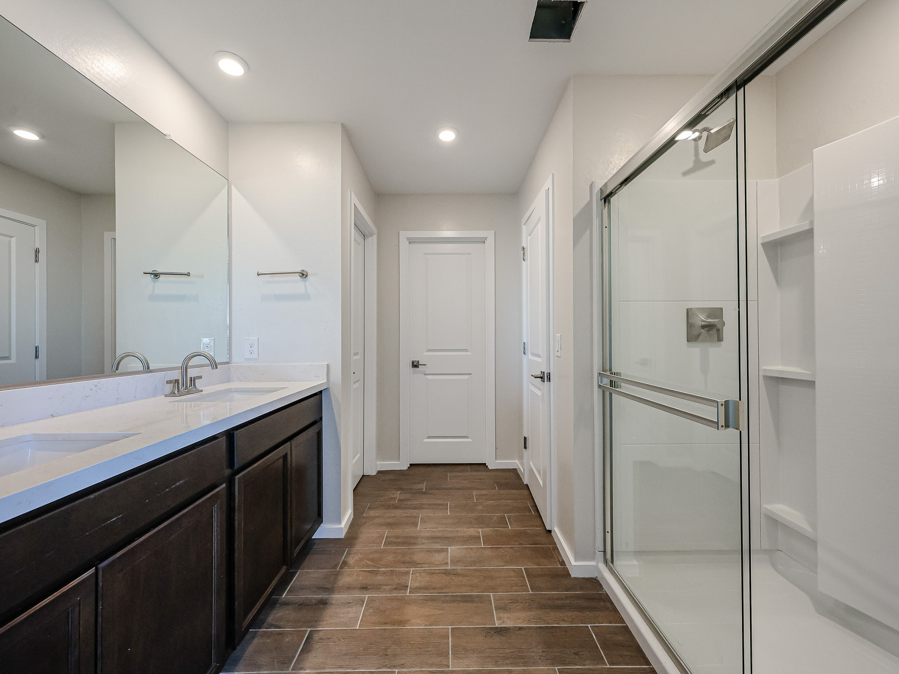 Modern bathroom with double sink vanity, large mirror, wood-look tile floor, glass-enclosed shower, and white walls with recessed lighting.