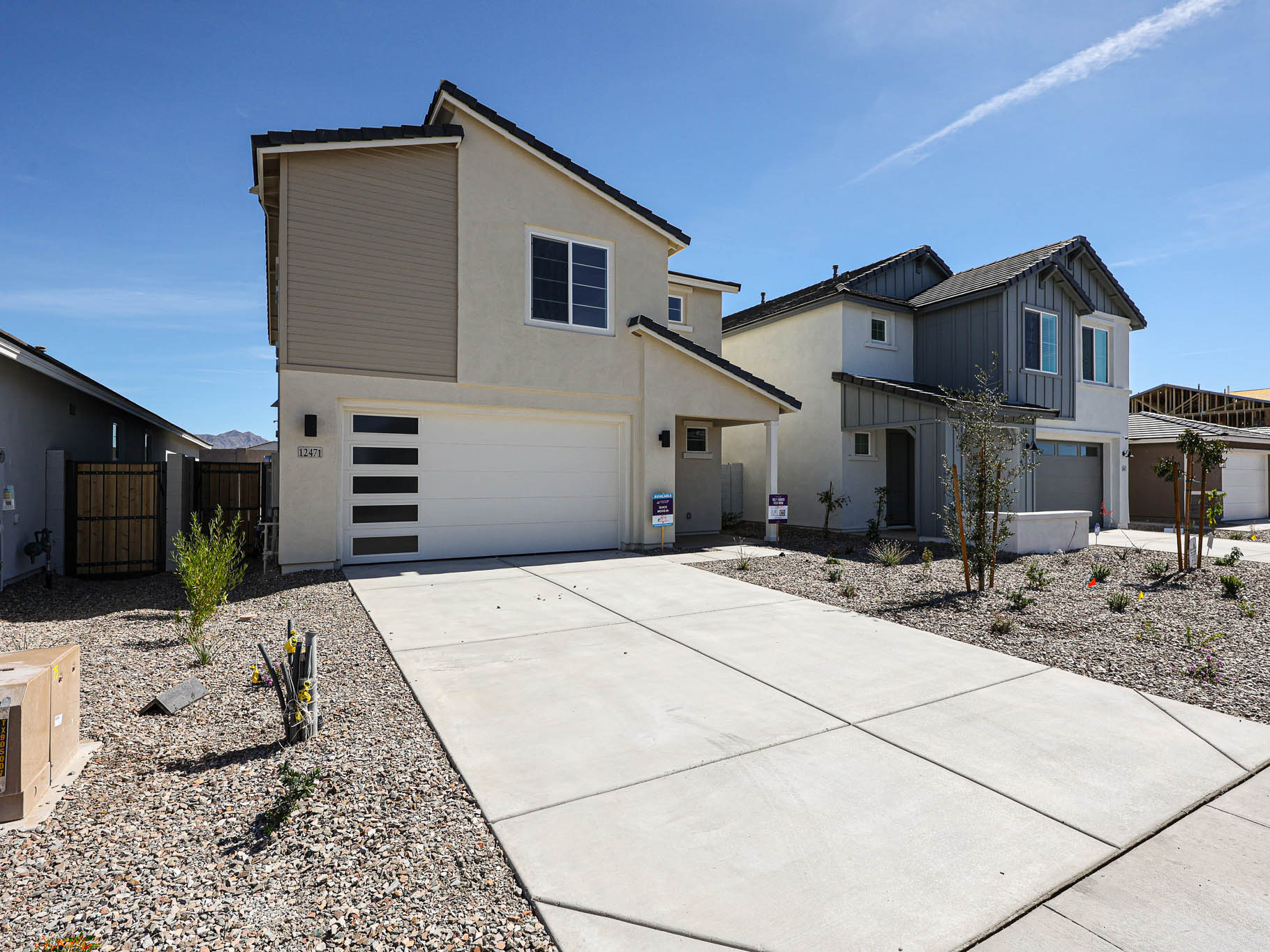 Two-story modern suburban house with a double garage, light-colored exterior, and a wide concrete driveway, surrounded by xeriscape landscaping. Other similar homes are nearby.