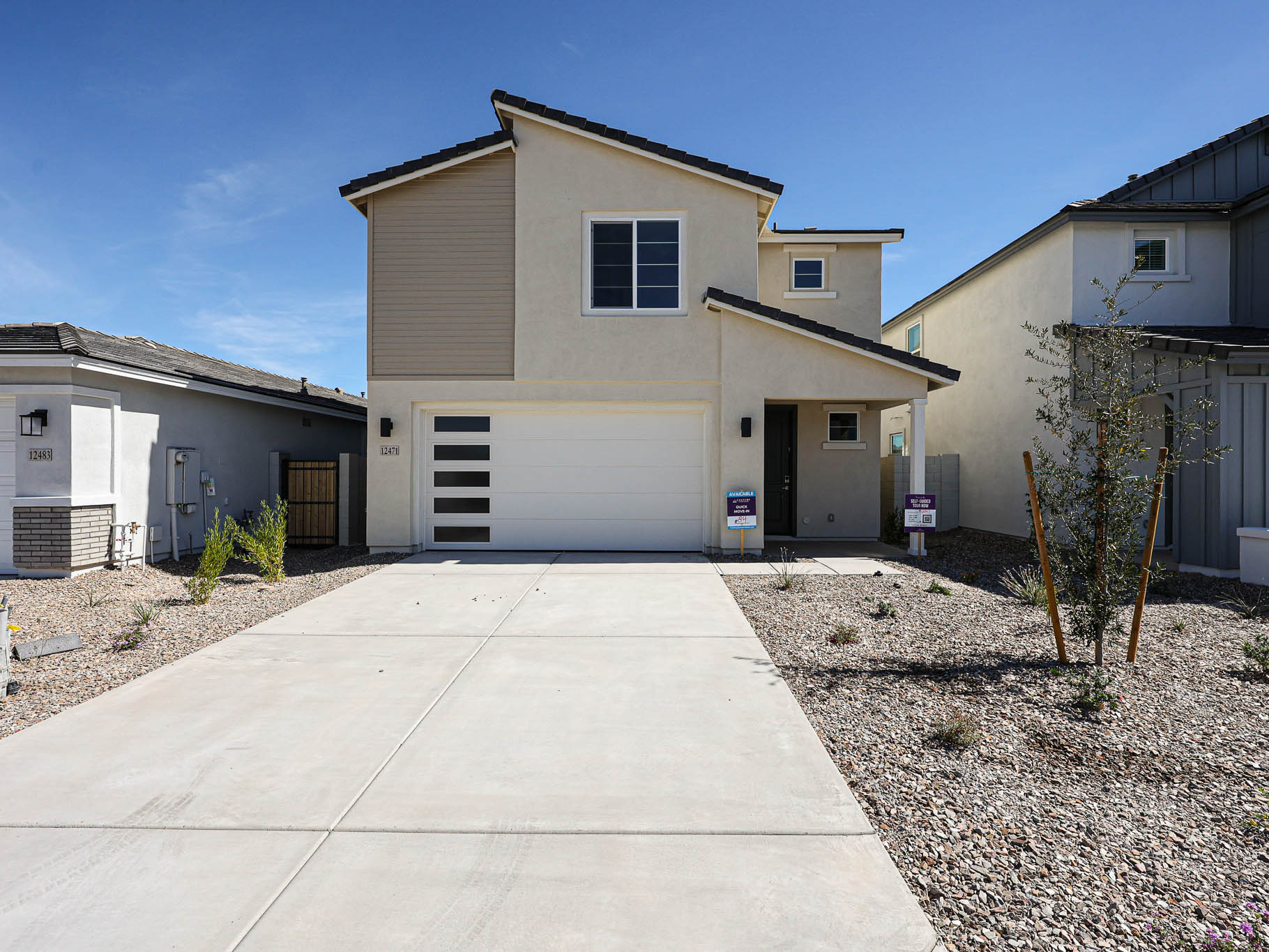 Two-story modern house with a beige exterior, garage, and a driveway. The front yard is landscaped with rocks and a few small plants. Signs are posted near the entrance.