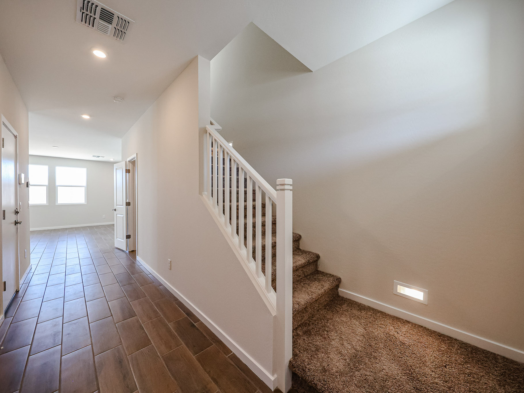A hallway with brown tile flooring leads to a carpeted staircase with a white railing and a room with large windows in the background.