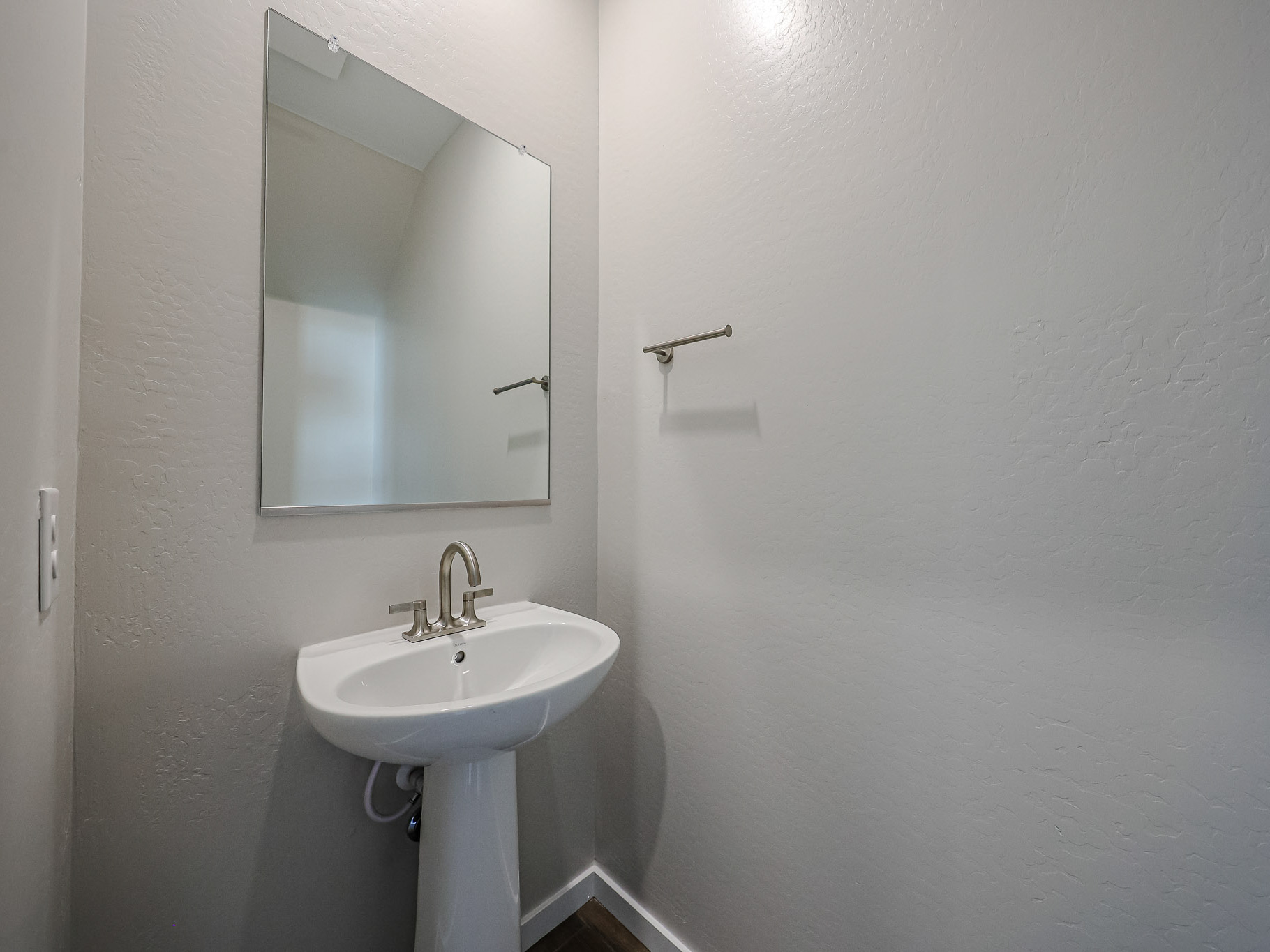 A small bathroom with a pedestal sink, a rectangular mirror above it, a wall-mounted towel bar, and light gray textured walls.