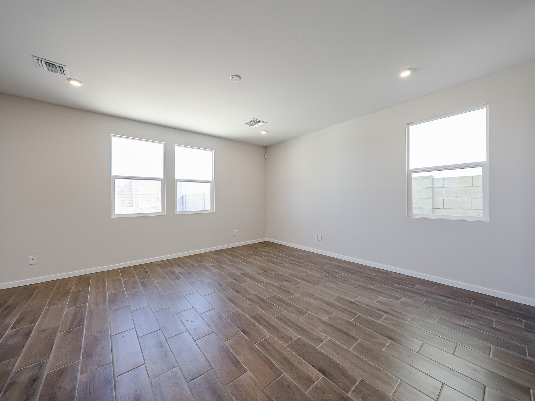 Empty room with light-colored walls, three windows, recessed ceiling lights, and wood-look tile flooring.