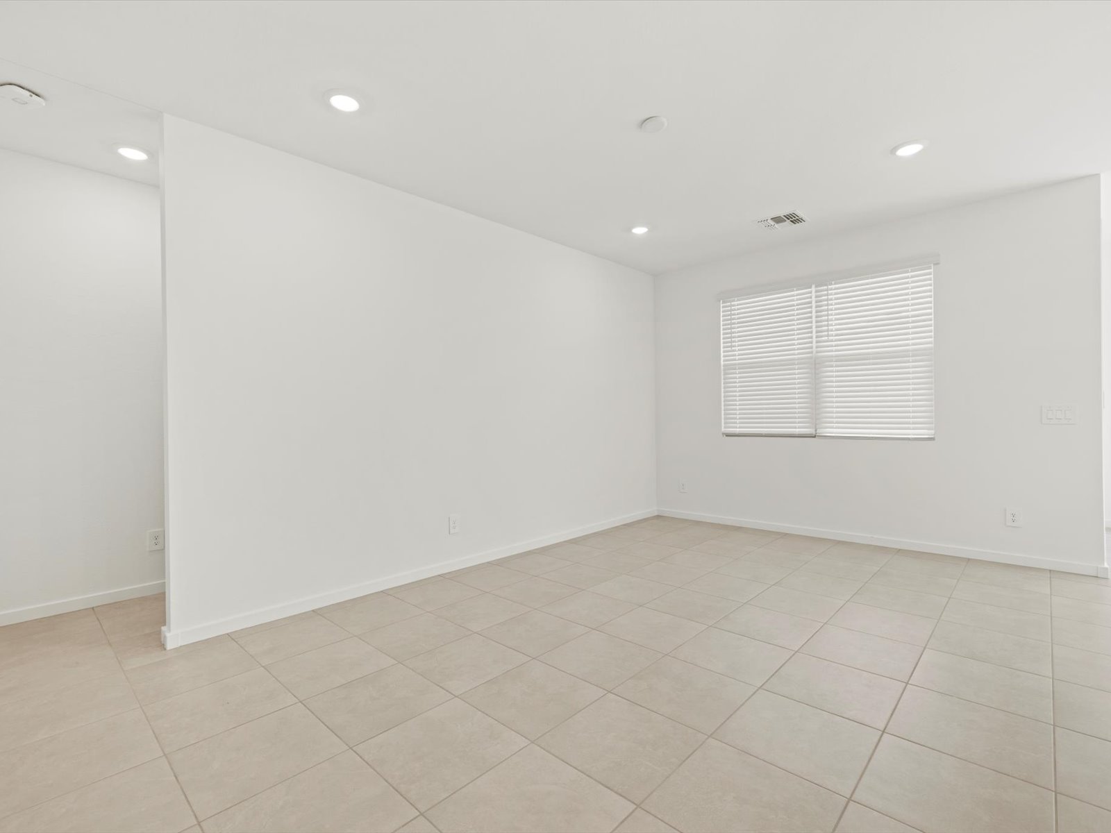 Empty room with white walls, beige ceramic tile floor, recessed ceiling lights, and a window with closed blinds letting in daylight.