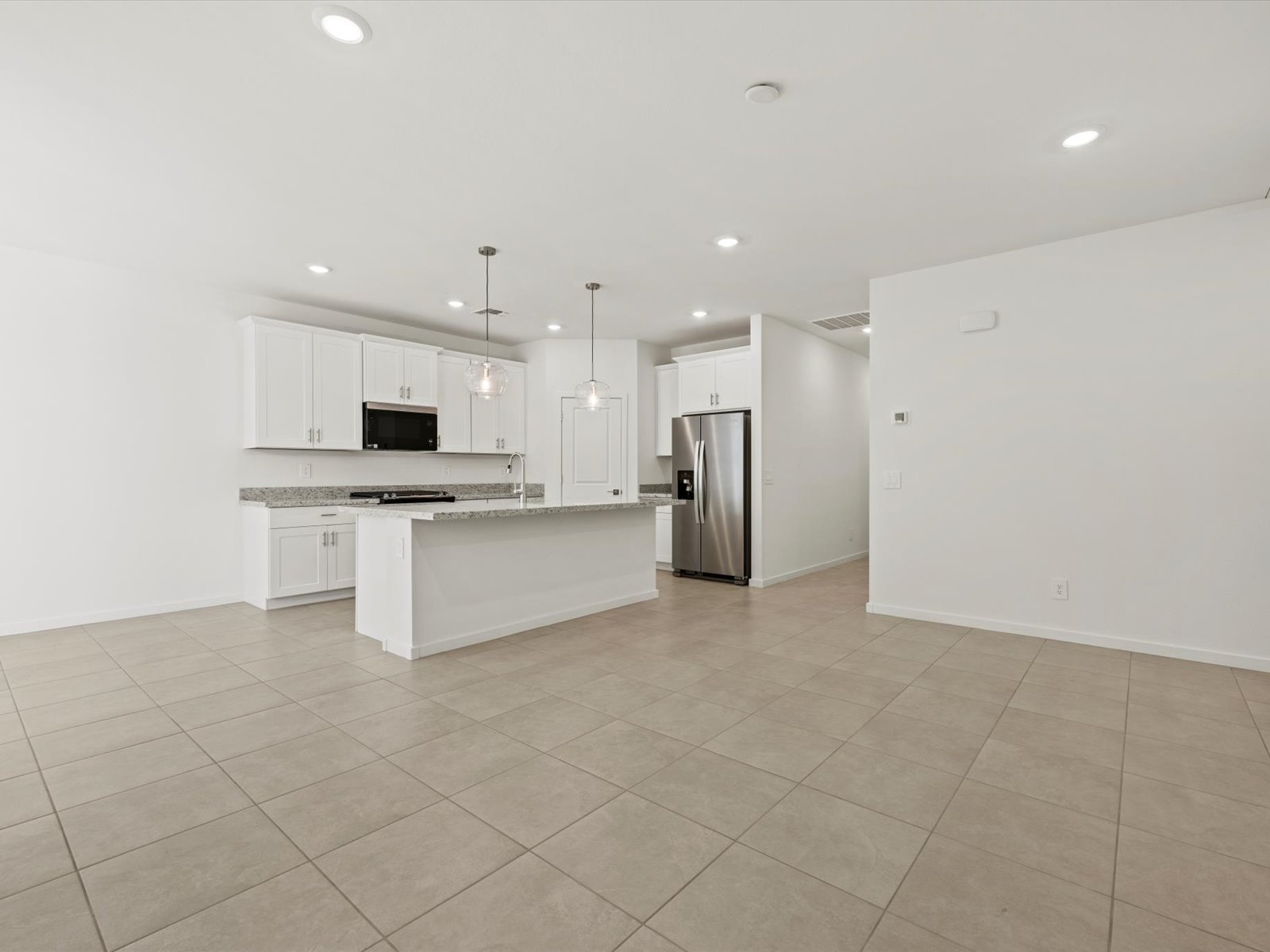 Spacious, modern kitchen with white cabinets, stainless steel appliances, and a central island, opening to a large tiled living area with recessed lighting.