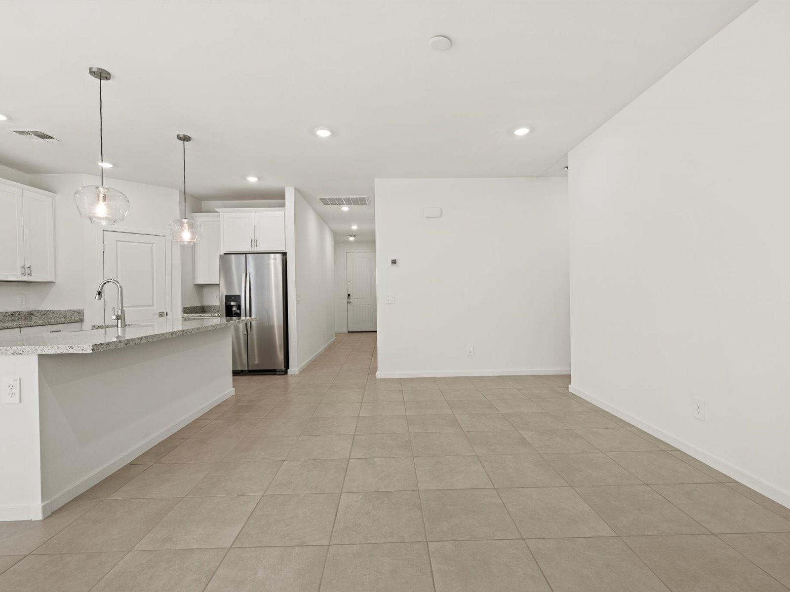 Modern open-concept kitchen and living area with tile flooring, white walls, stainless steel appliances, and pendant lighting over a granite countertop island.