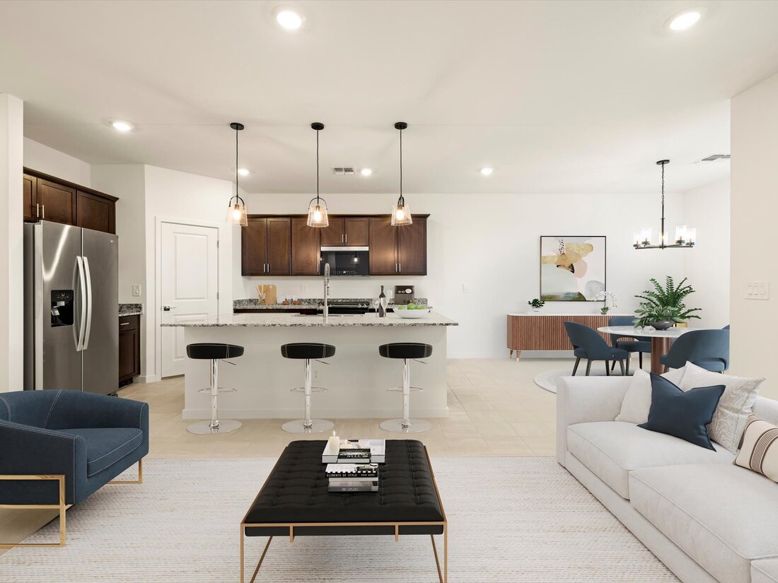 Open-concept kitchen and living area with modern furniture, a large island with barstools, stainless steel appliances, and pendant lighting. Dining table and artwork are visible in the background.