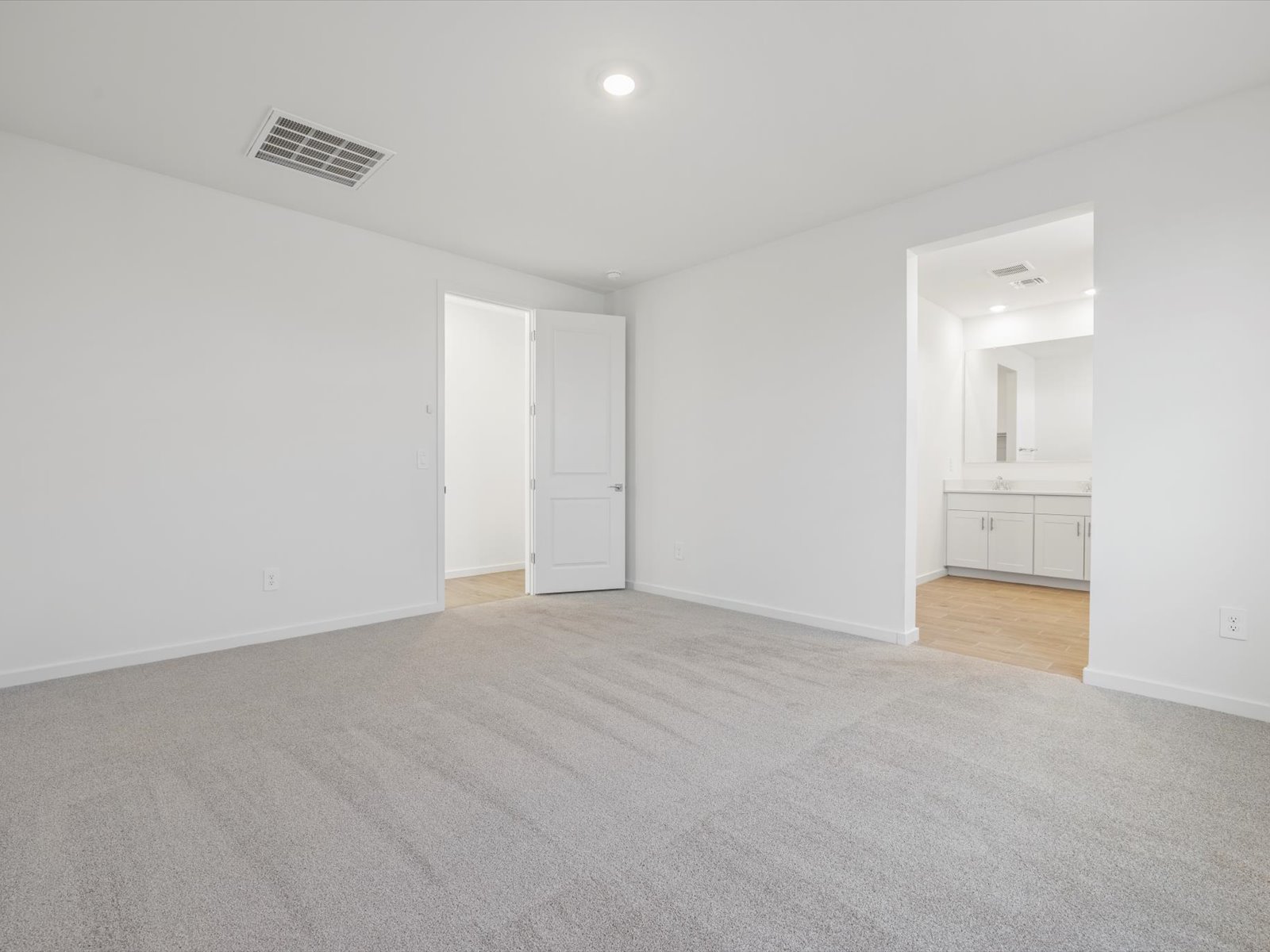 Unfurnished bedroom with light gray carpet, white walls, a closed door, and an open doorway leading to a bathroom with a double-sink vanity.