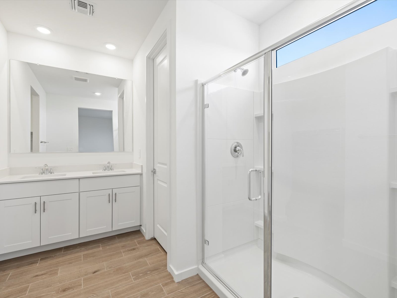 Modern bathroom with a double-sink vanity, large mirror, glass-enclosed shower, white walls, and wood-look tile flooring.