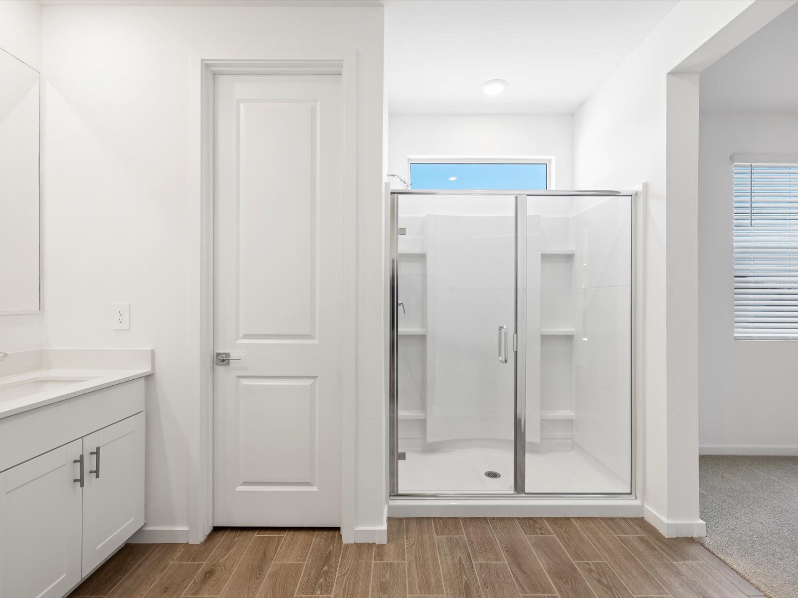 Modern bathroom with a glass shower enclosure, white cabinets with a sink, wood-look tile flooring, and a window providing natural light.