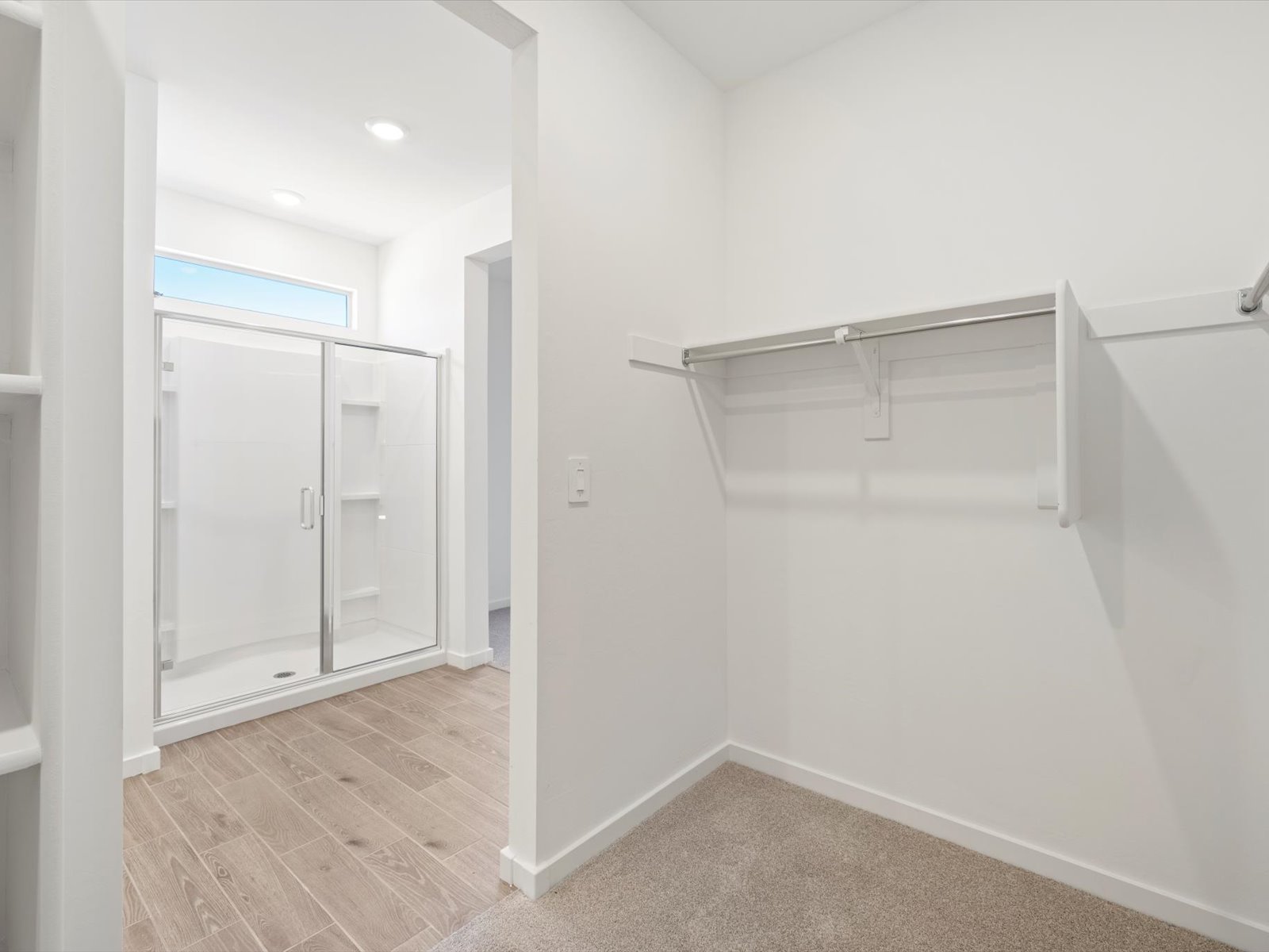 A walk-in closet with carpet flooring is next to a bathroom area featuring a glass-enclosed shower and tile floor.