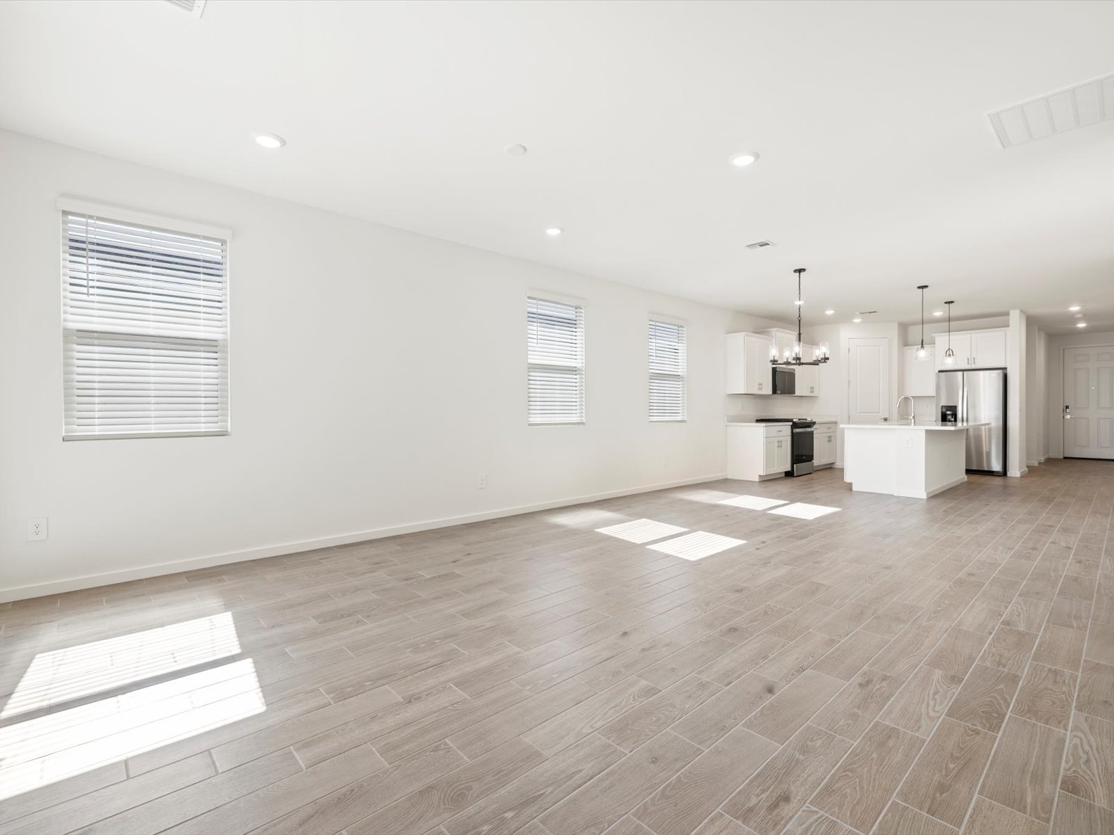 Spacious, empty open-concept living area with wood-look tile flooring, white walls, multiple windows, and a modern kitchen featuring white cabinets and an island with pendant lights.