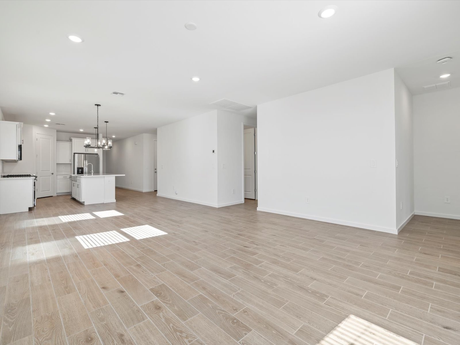 Empty modern open-concept room with wood-look tile flooring, white walls, recessed lighting, and a view of the kitchen with an island and pendant lights in the background.