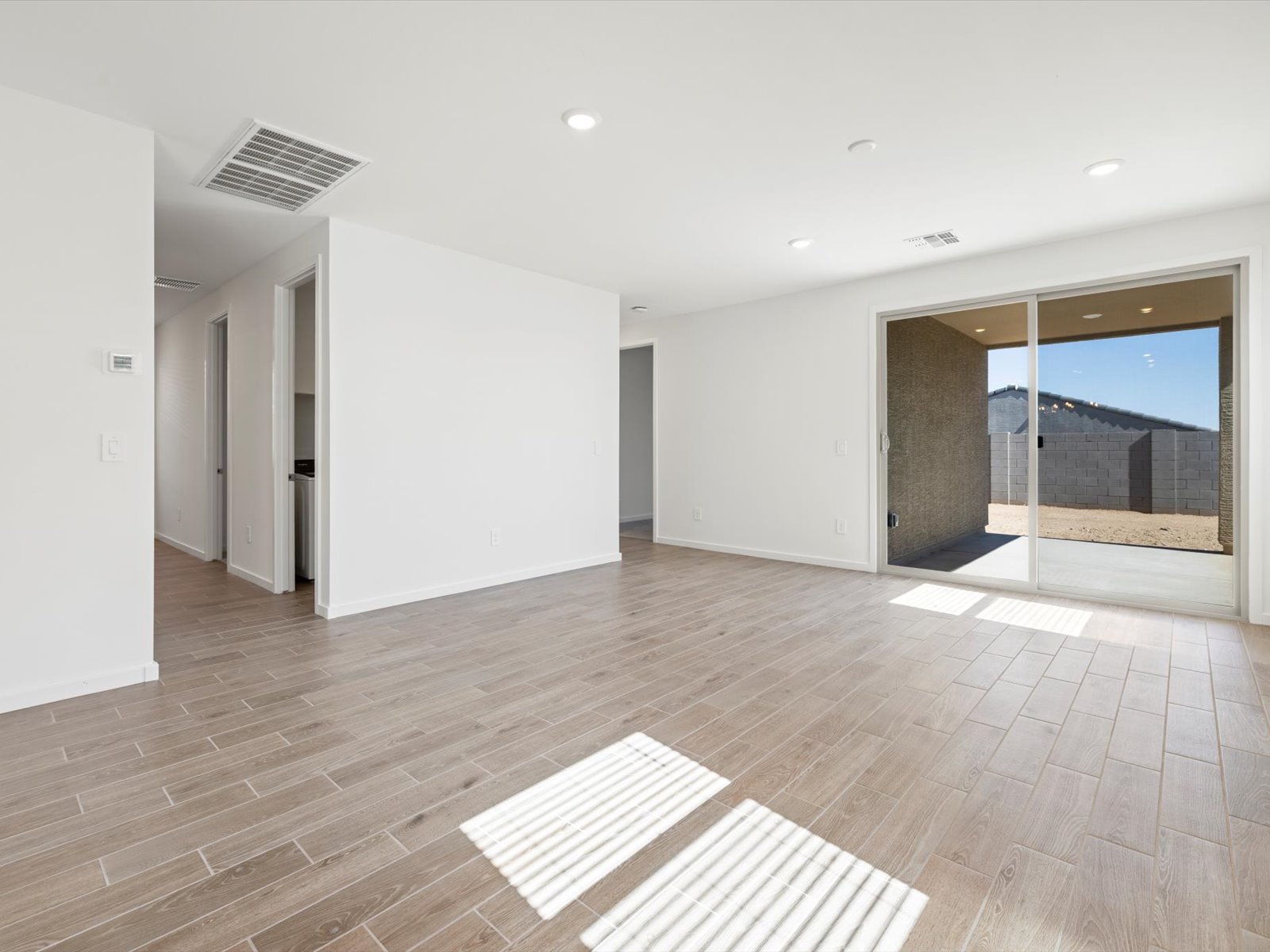 Empty modern living room with light wood flooring, white walls, recessed lighting, sliding glass doors, and view of a backyard with a block fence.