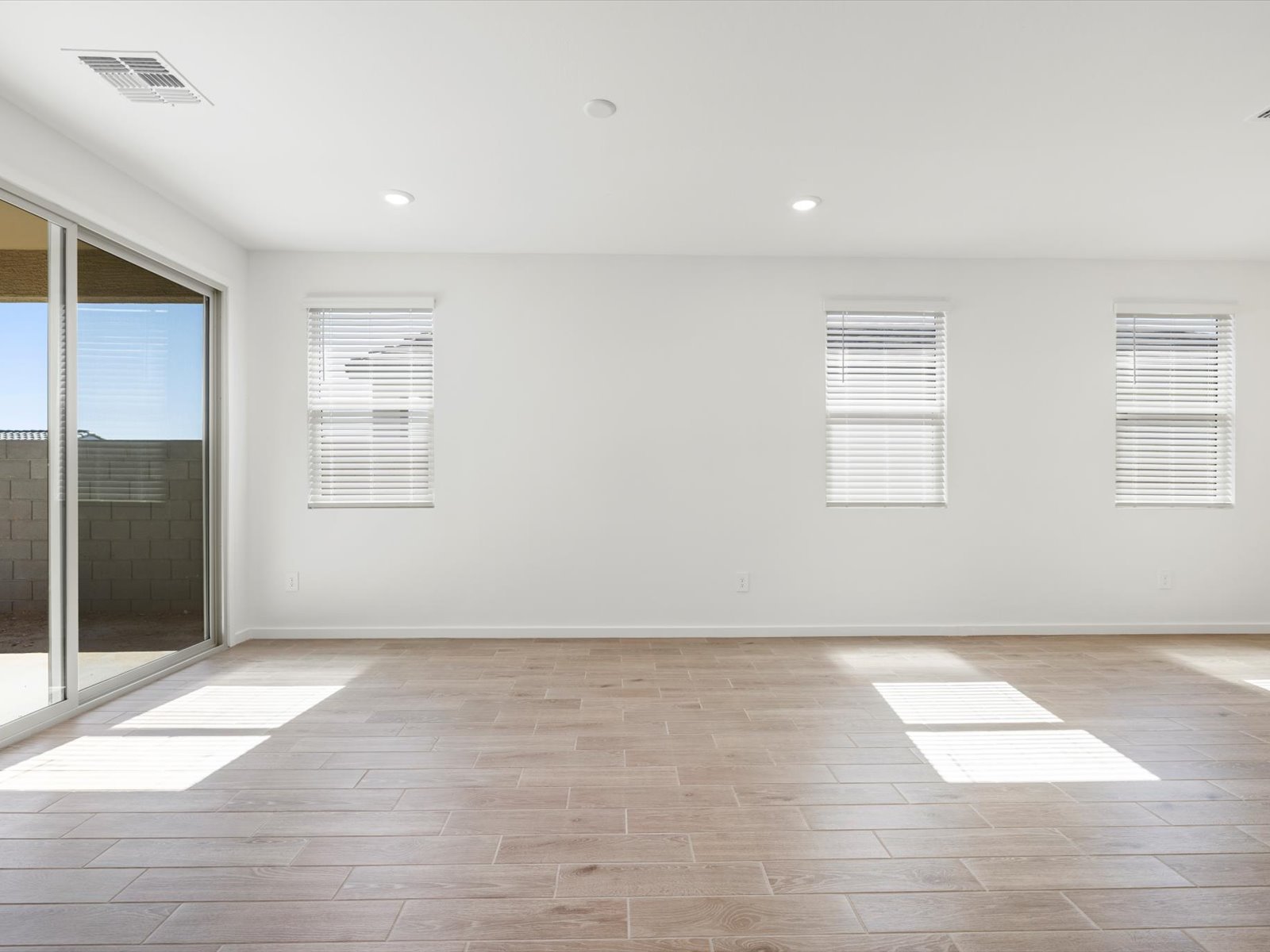 Empty room with light wood flooring, three windows with white blinds, recessed lights, and a sliding glass door leading to a patio and fenced backyard.