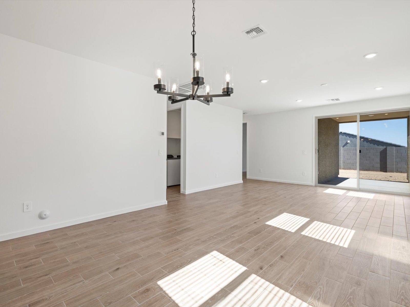 Empty room with wood-look tile flooring, white walls, a black modern chandelier, and large sliding glass doors leading to a fenced backyard. Sunlight streams through the doors onto the floor.