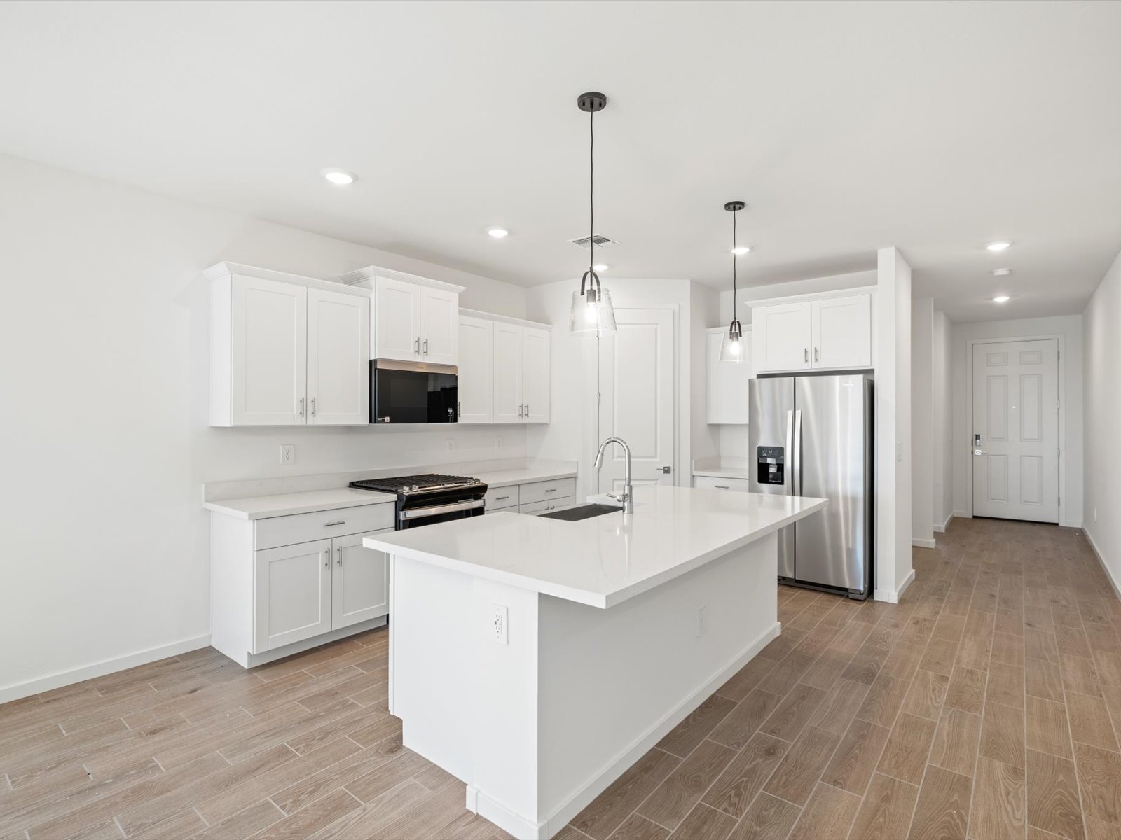Modern kitchen with white cabinets, stainless steel appliances, an island with sink, pendant lights, and wood-look tile flooring. Hallway and door visible in the background.