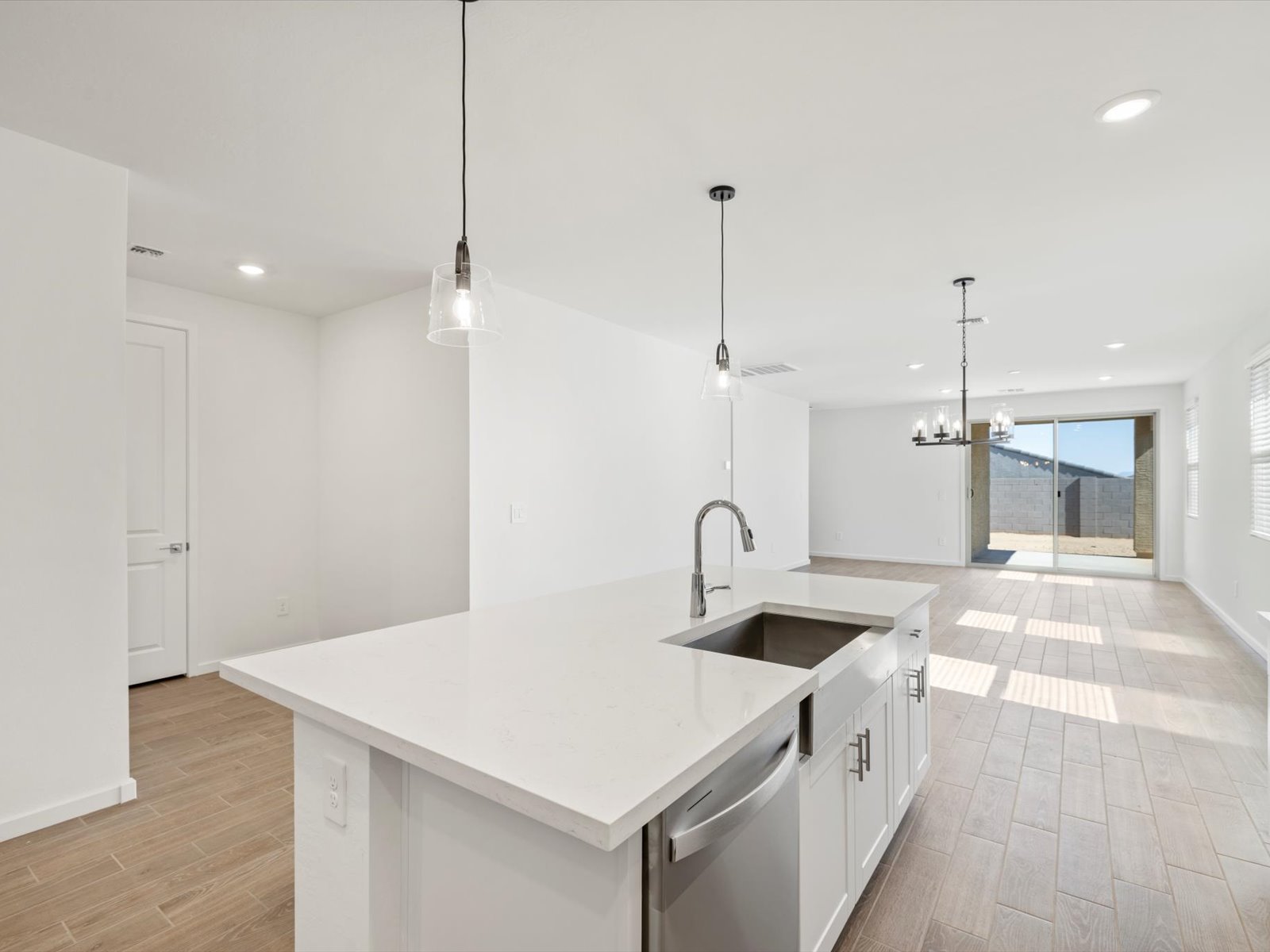 Modern kitchen with white island, stainless steel appliances, pendant lights, and open layout leading to a bright living area with large windows and wood-look tile flooring.