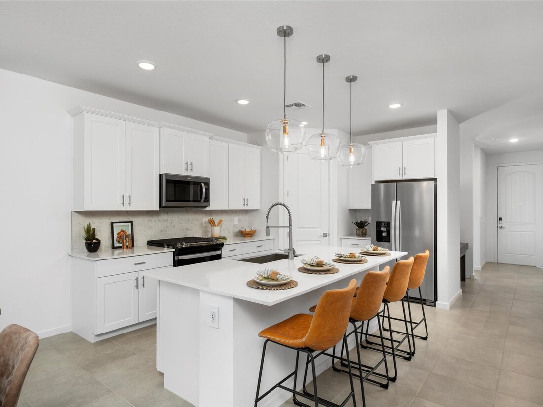 Modern kitchen with white cabinets, stainless steel appliances, a large island with sink, and four tan barstools. Three pendant lights hang above the island, and the room is well-lit.