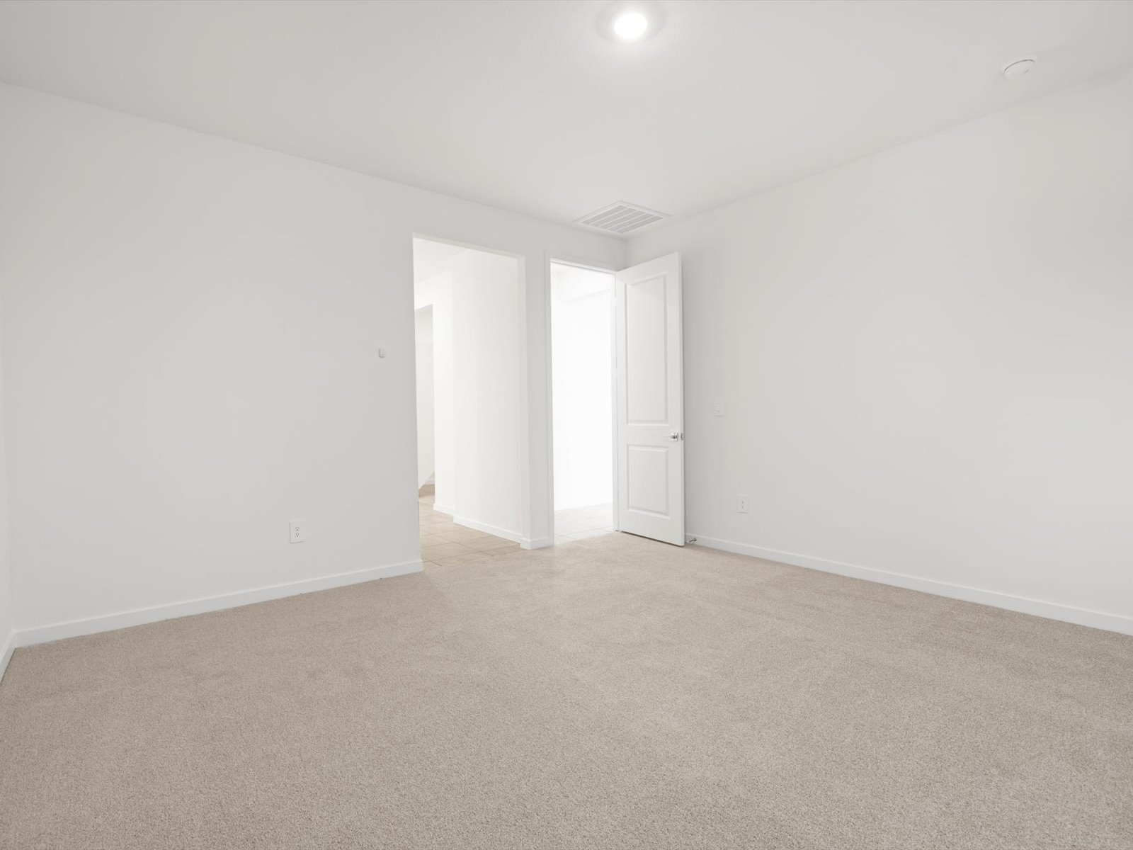 Empty room with beige carpet, white walls, a ceiling light, and an open door leading to a brighter hallway.