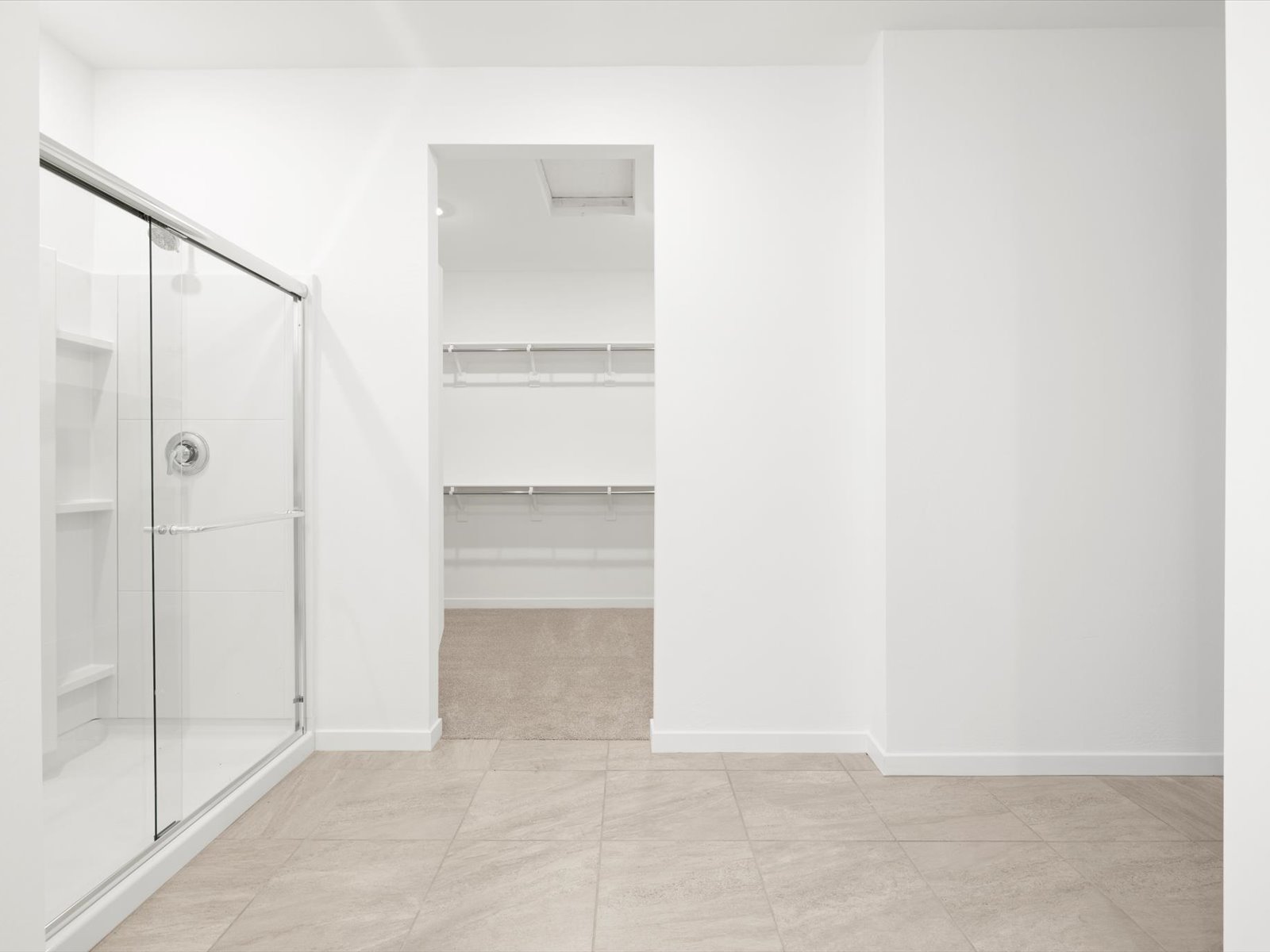 Minimalist bathroom with a glass shower on the left, beige tiled floor, and an open doorway leading to an empty walk-in closet with shelves and hanging rods.