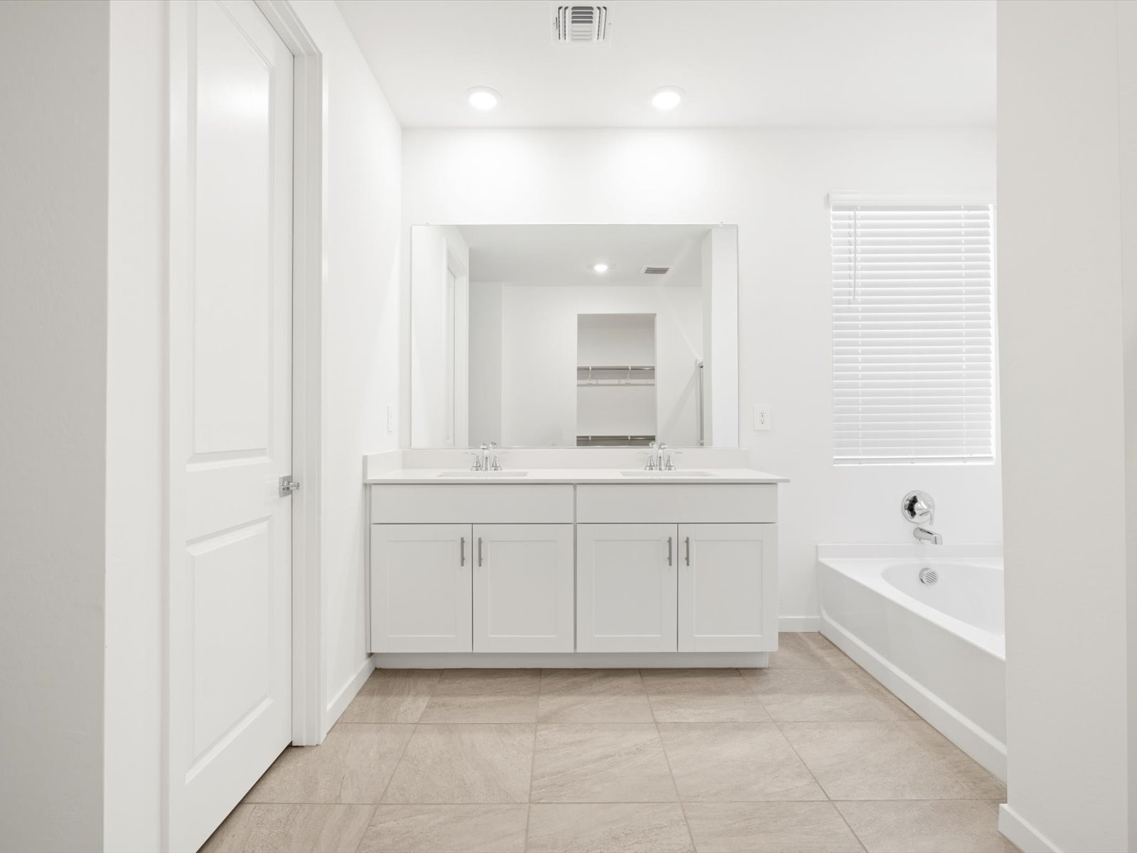 Modern bathroom with white cabinets, double sinks, large mirror, bathtub, tiled floor, and a window with closed blinds.