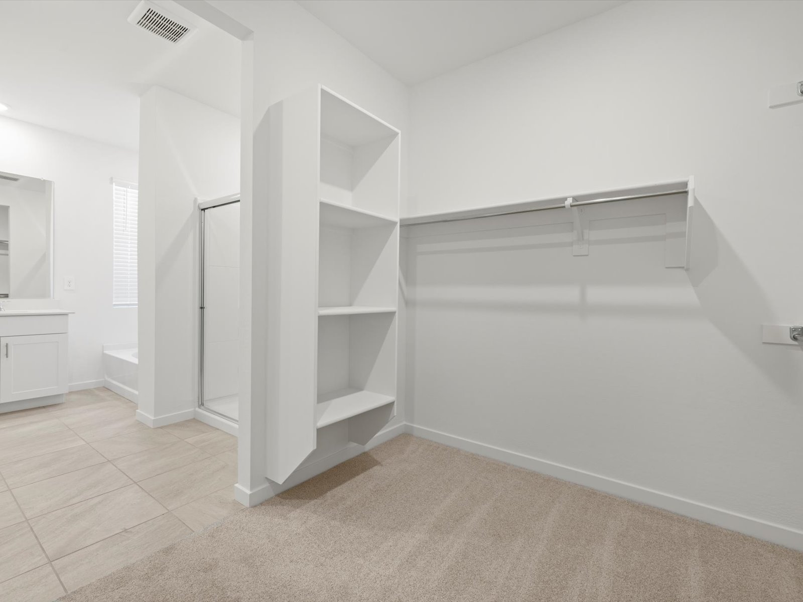 Spacious walk-in closet with built-in shelves, hanging rods, and beige carpet, adjacent to a bathroom with tile flooring and a shower.