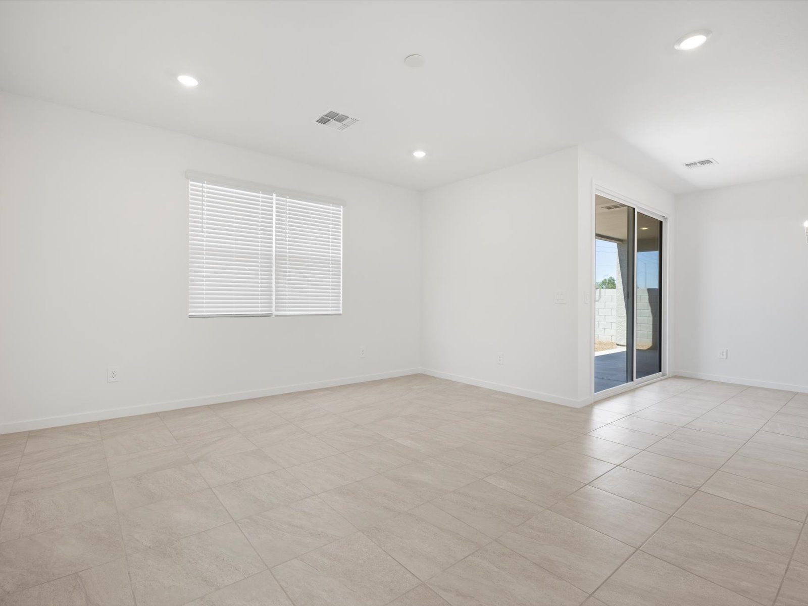 Empty room with beige tiled floor, white walls, window with blinds, recessed ceiling lights, and sliding glass door leading to an outdoor patio.