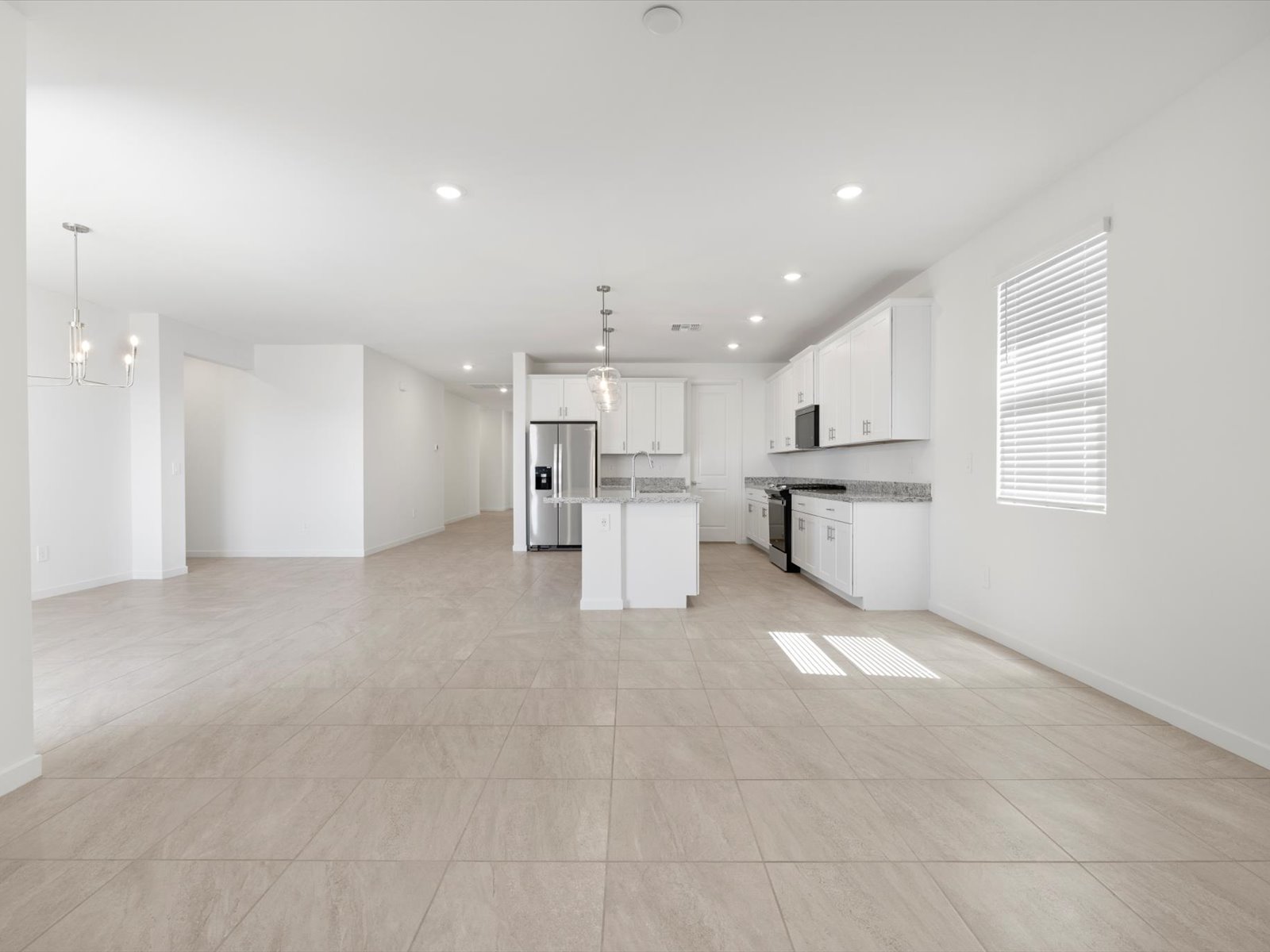 Spacious, empty modern kitchen and dining area with white cabinets, stainless steel appliances, tiled floor, and large windows letting in natural light.