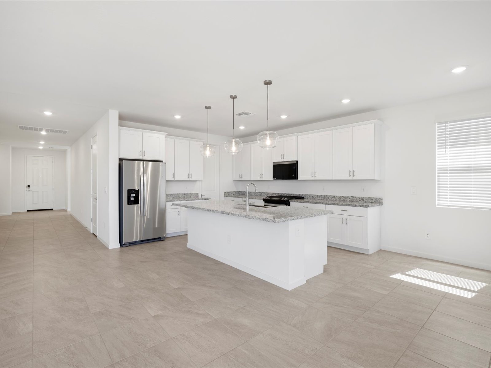 Modern kitchen with white cabinets, stainless steel appliances, granite countertops, island with sink, three pendant lights, and tiled floor in a bright, open space.