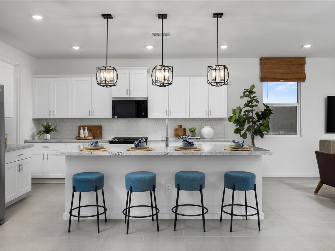 Modern kitchen with white cabinets, an island with four blue stools, pendant lights overhead, and place settings on the counter. A plant and window with brown shade are visible.