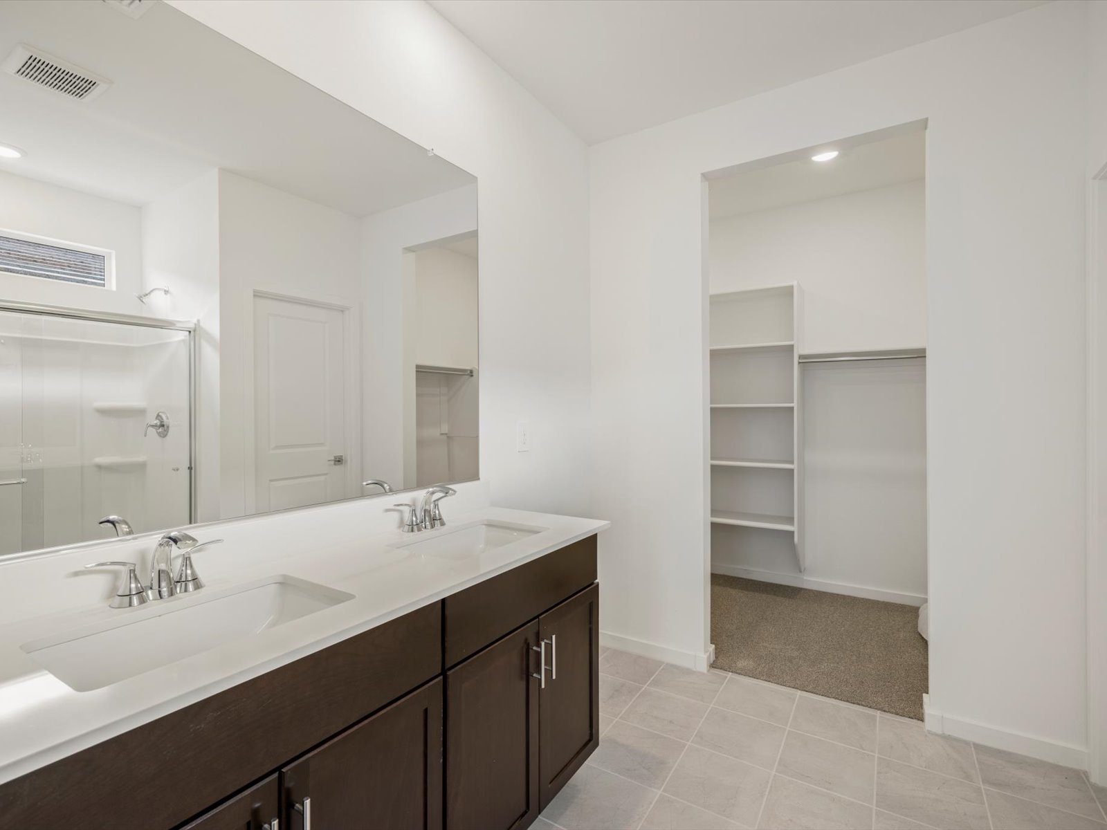 Modern bathroom with double sink vanity, large mirror, walk-in shower with glass door, and adjacent walk-in closet with built-in shelves and hanging rods.
