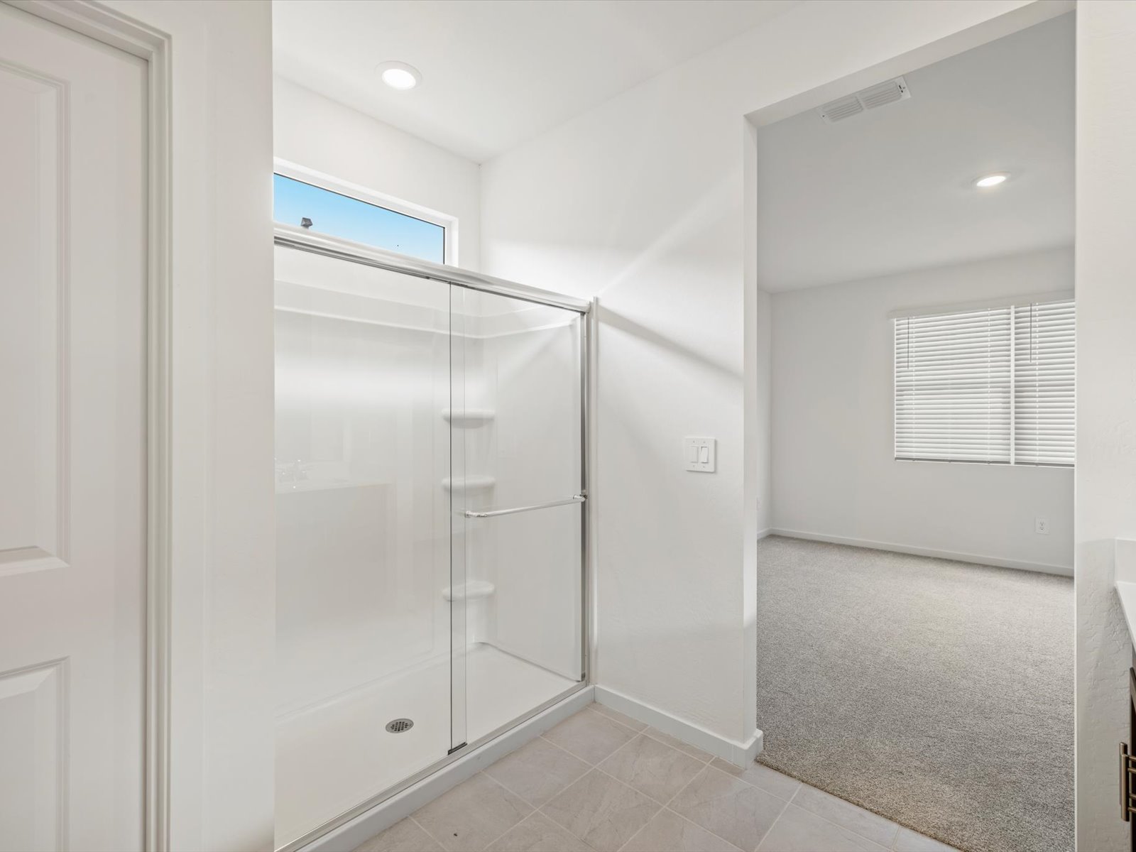 A bathroom with a glass shower enclosure and tiled floor, connected to a carpeted bedroom with a large window and closed blinds.