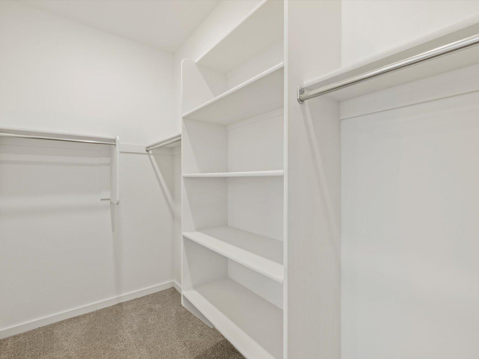 Empty walk-in closet with white shelves, hanging rods, and beige carpet flooring. The closet has white walls and is well-lit.