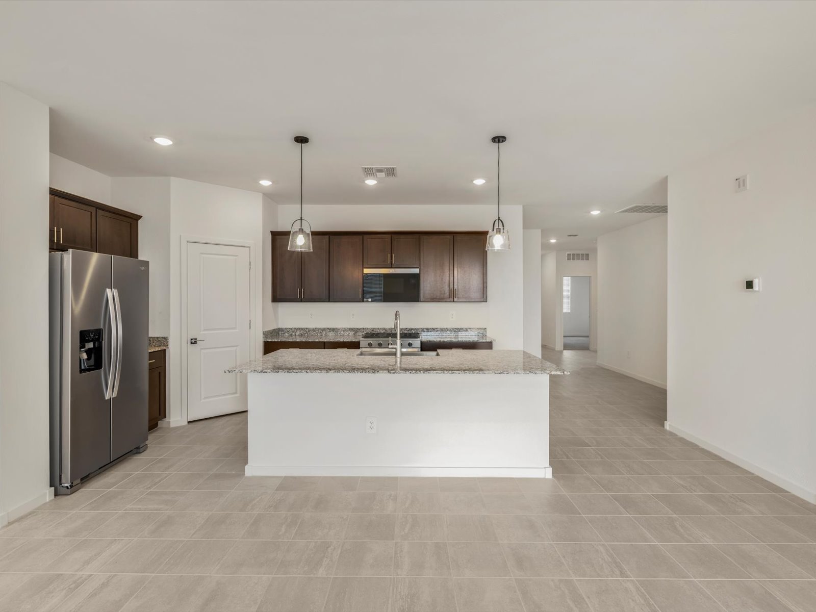 Modern kitchen with stainless steel refrigerator, dark wood cabinets, granite island with sink, pendant lights, and beige tile flooring. White walls and open floor plan.