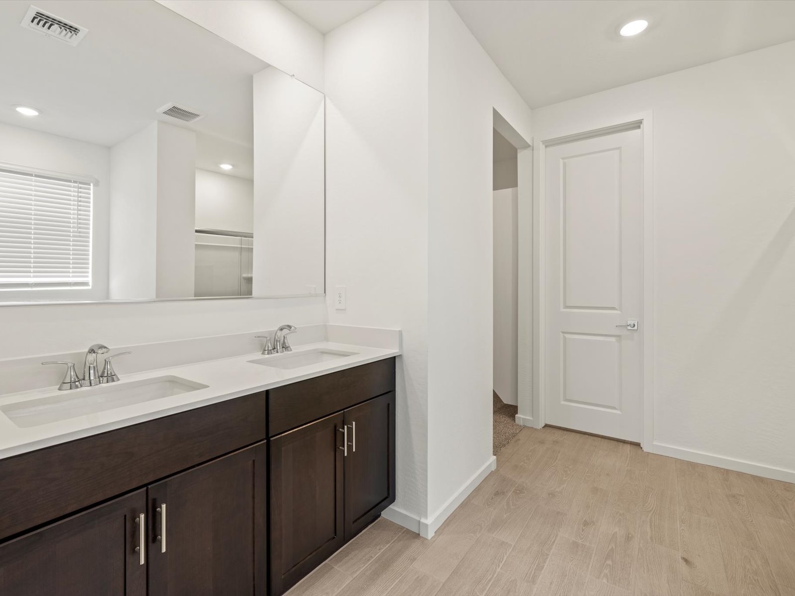 Modern bathroom with double sinks, dark wood vanity, large mirror, light wood flooring, and white walls. Door and window visible in the background.
