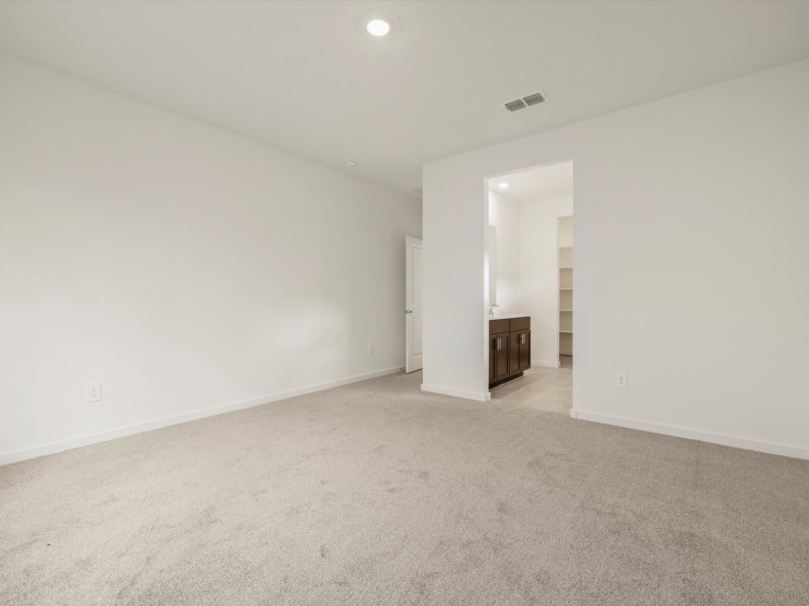 Empty room with beige carpet, white walls, a doorway leading to a bathroom with a vanity, and built-in shelving visible in the background.