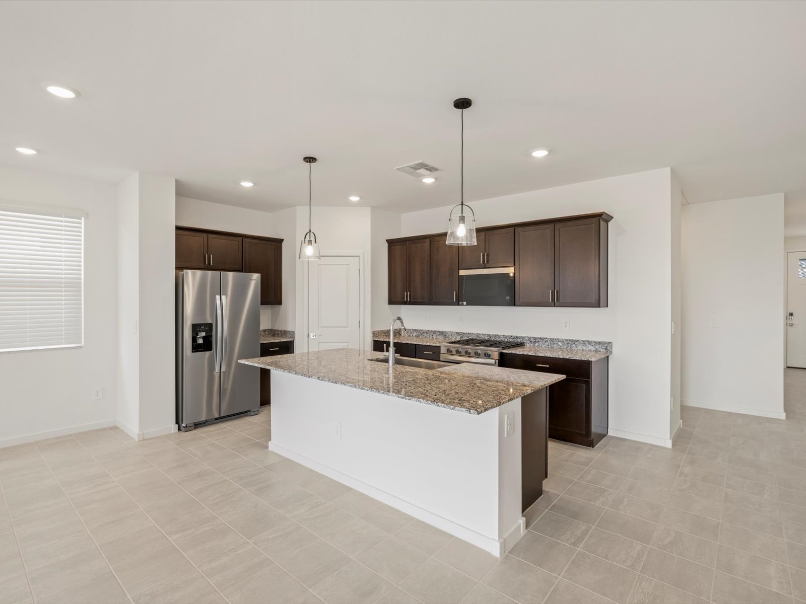 Modern kitchen with stainless steel appliances, dark wood cabinets, granite countertops, center island with sink, and pendant lights; light tile flooring and white walls throughout.