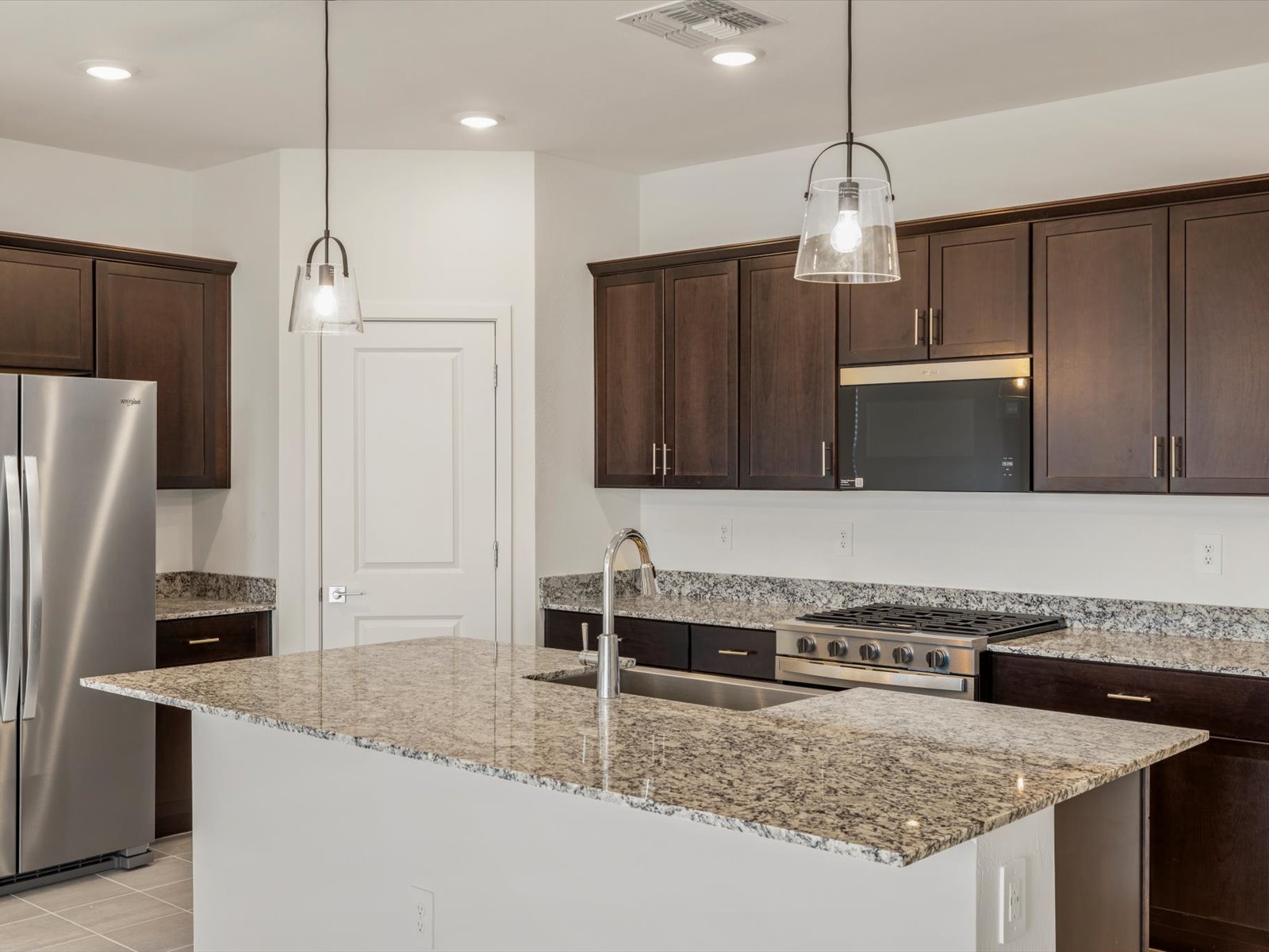Modern kitchen with dark wood cabinets, stainless steel appliances, granite countertops, pendant lighting, and an island with a built-in sink and faucet.