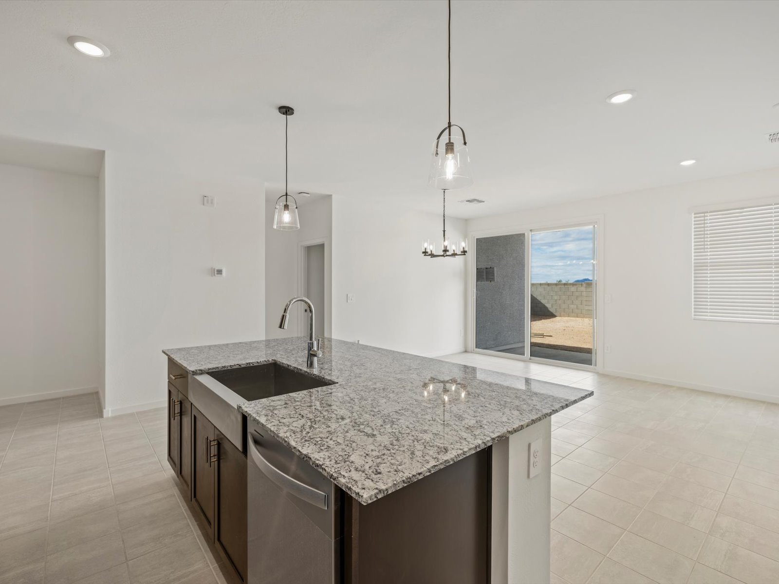 Modern kitchen with granite island, sink, pendant lights, and open living area; large windows and sliding glass door offer a view of a fenced backyard with a clear sky.