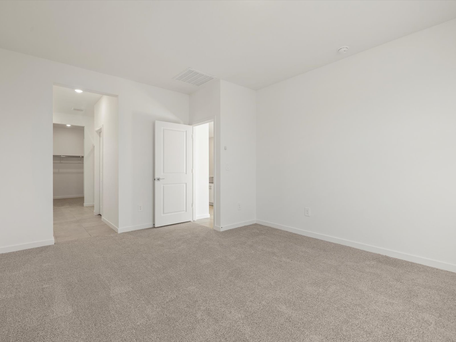 Empty bedroom with beige carpet, white walls, an open door leading to a bathroom, and a walk-in closet visible through another doorway. Room is well-lit with natural and ceiling light.
