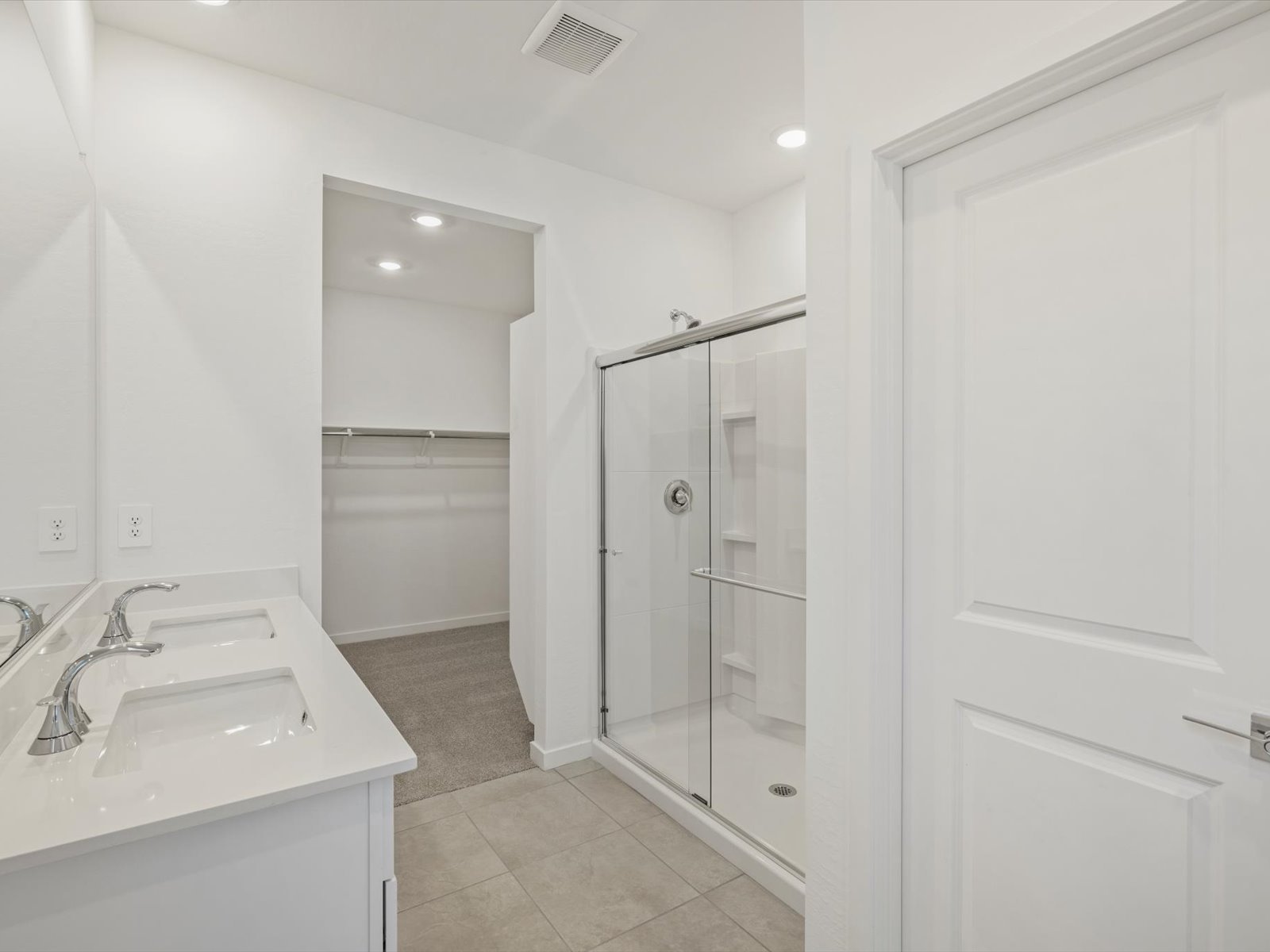 Modern bathroom with double sinks, large mirror, glass-enclosed shower, and doorway leading to a walk-in closet with built-in shelving. Floors are tiled; walls and cabinets are white.