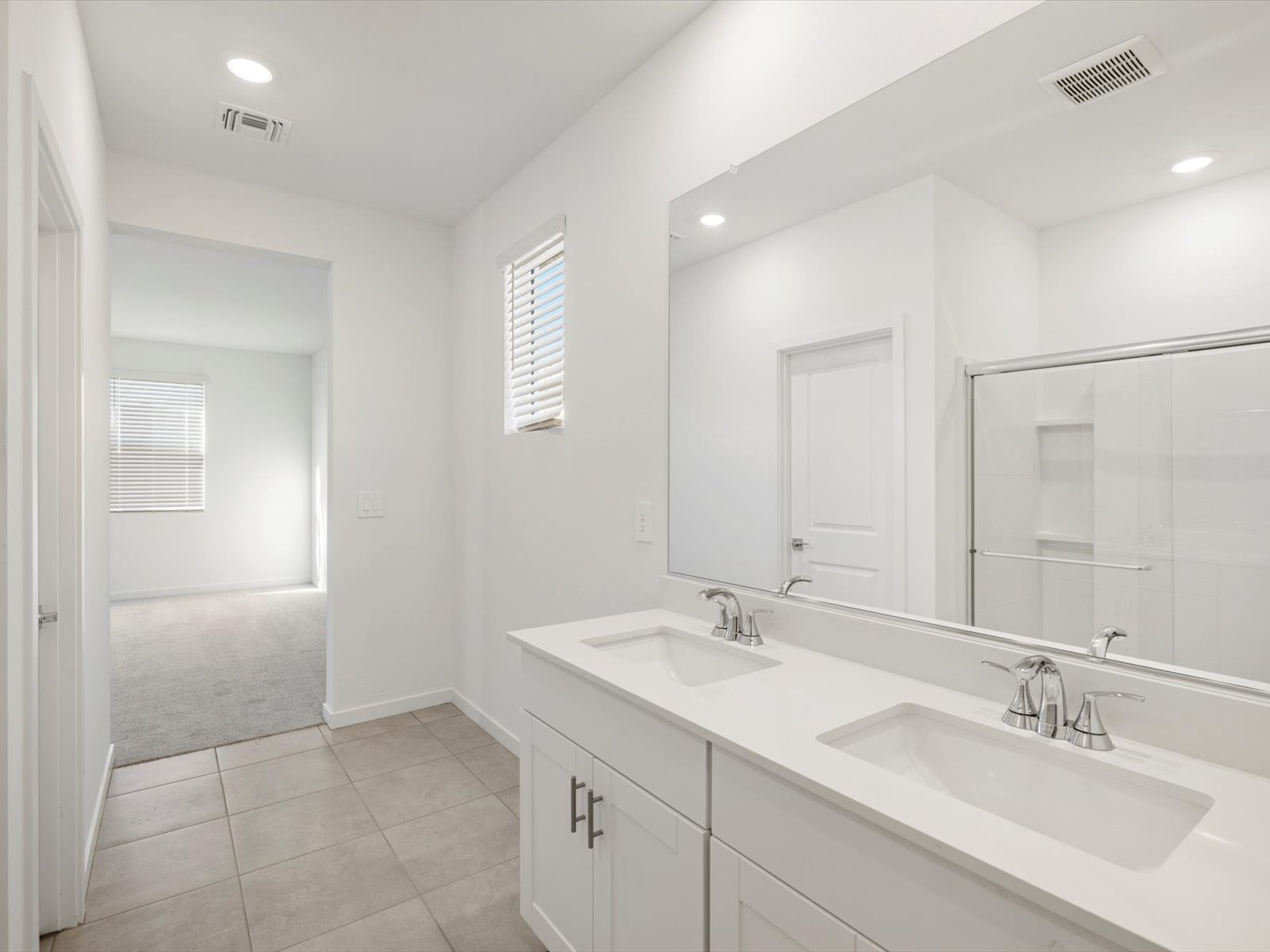 Modern bathroom with double sinks, white cabinets, a large mirror, tiled floor, and a glass shower; a doorway leads to a carpeted room with windows.