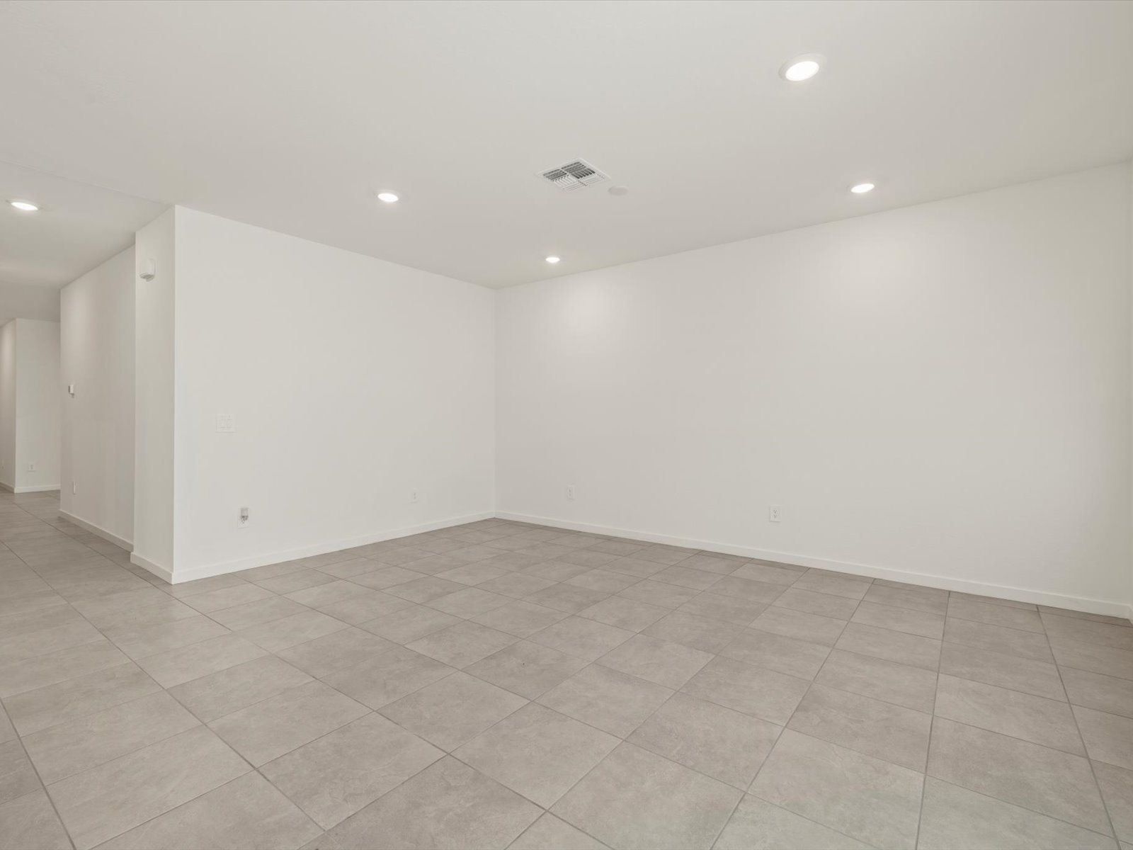 Empty room with light gray floor tiles, white walls, recessed ceiling lights, and a central air vent; hallway visible to the left.