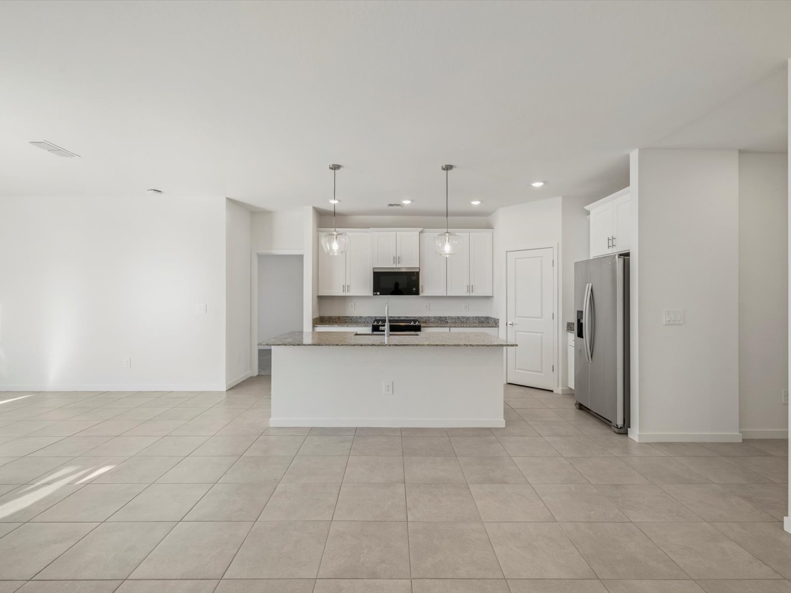 Modern kitchen with white cabinets, stainless steel appliances, granite countertops, pendant lights, and an island, opening to a spacious tiled living area.