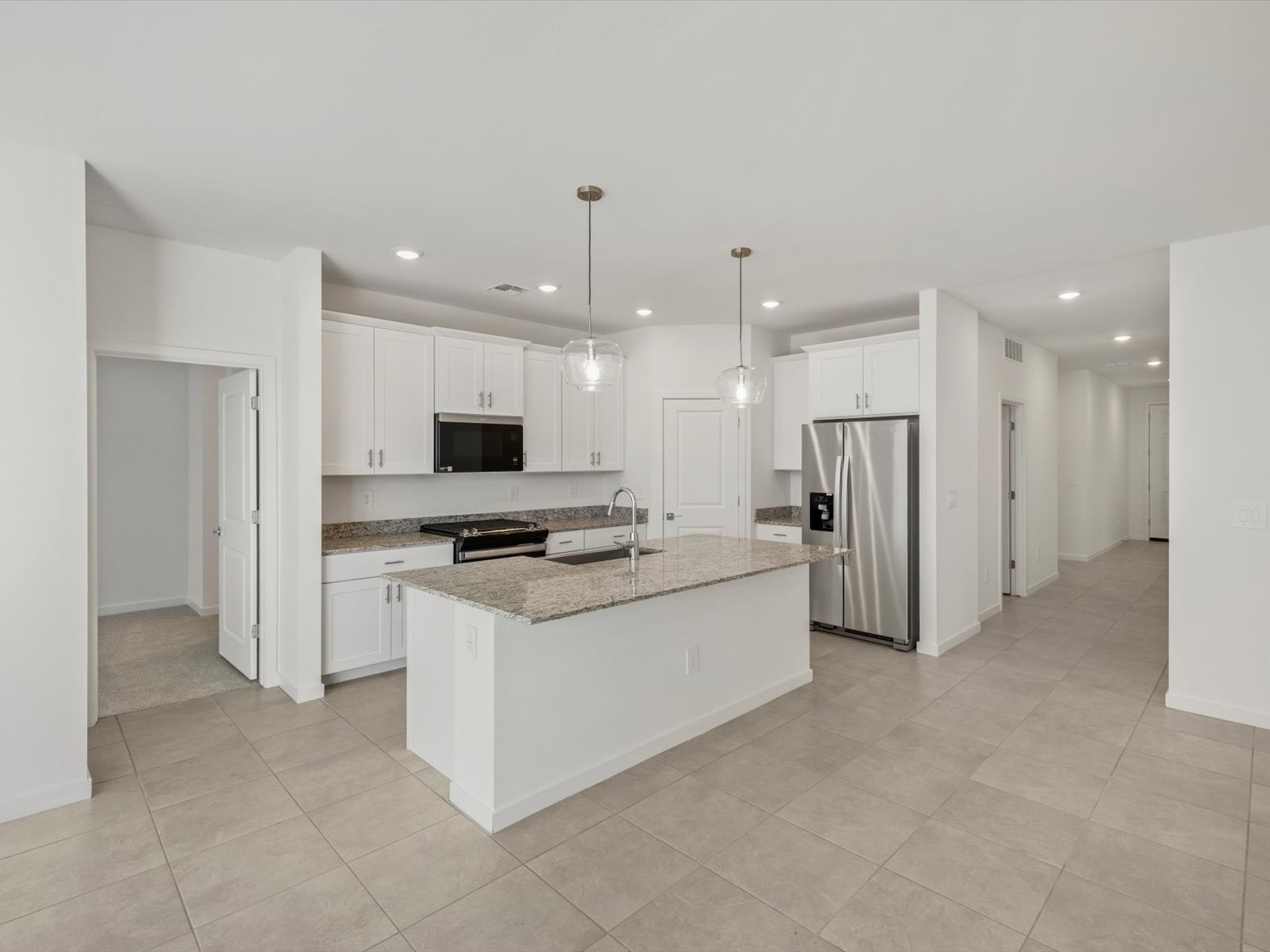 Modern kitchen with white cabinets, granite island, stainless steel appliances, pendant lights, and tiled floor, adjacent to a hallway and open doorway.