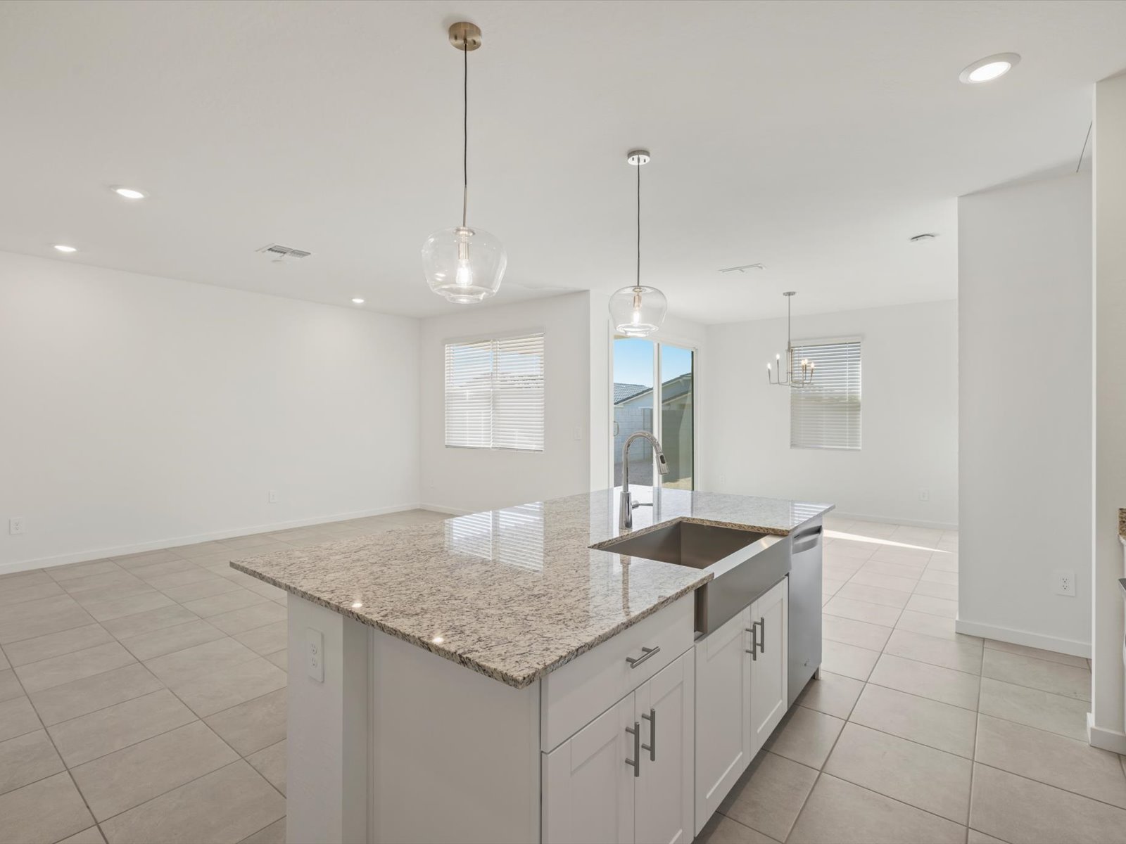 Modern kitchen with granite island, stainless steel sink, pendant lights, tile flooring, and open dining and living area with large windows and sliding glass door.