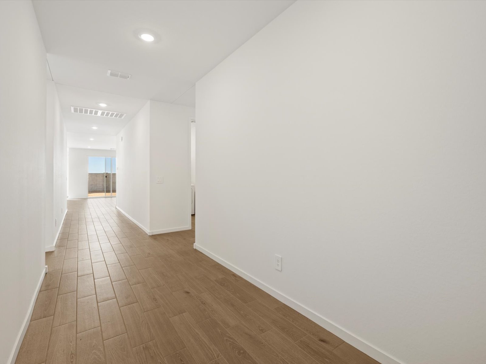 A long hallway with white walls, wood-look tile flooring, ceiling lights, and a view of a sliding glass door at the far end.